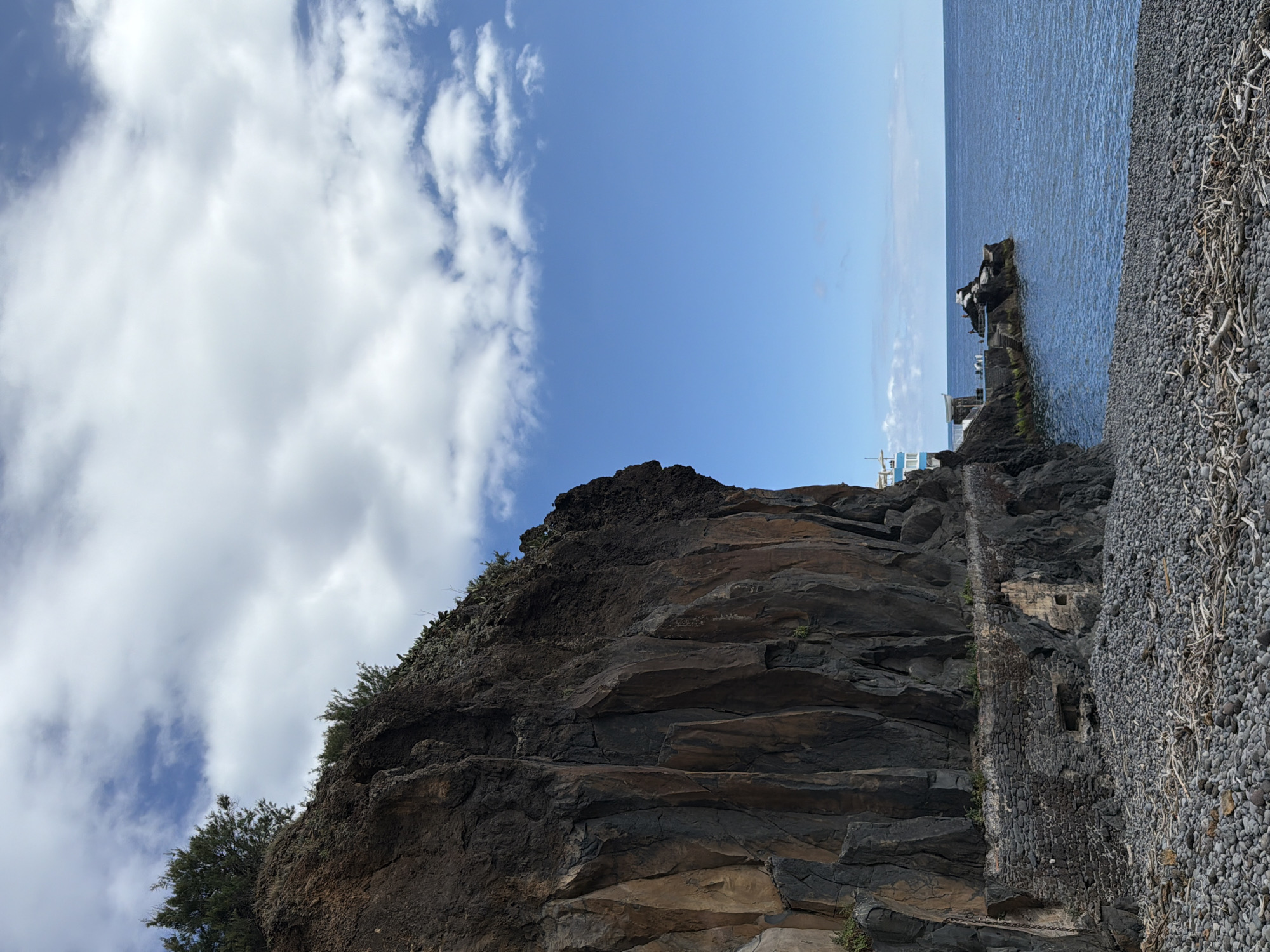 This outdoor, rotated, eye-level shot shows a coastal scene in Funchal, Portugal. The sky is partly cloudy, with patches of blue. A cliff dominates the left side of the frame, showing stratified rock layers. Vegetation clings to the cliff face.
To the right, a steep bank covered in pebbles meets the ocean. A narrow, dilapidated stone structure runs along the cliff face and the pebble bank, appearing to be an old retaining wall or part of a former coastal fortification. A concrete structure is on a rocky outcrop at the edge of the sea with a few people on it. The horizon is visible in the upper center of the frame, separating the sky from the ocean.