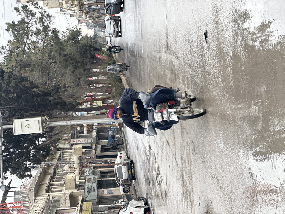 This image depicts a street scene within Quetta City Tehsil, Pakistan, on what appears to be an overcast day. The street surface is wet with standing water and reflections, indicating recent rain.
In the foreground, a male individual is operating a silver/chrome motorcycle, likely a Honda model given its design. He is wearing a dark blue sweater with a yellow "W" logo and "Sun Jan" text, blue jeans, a pink and blue knit cap, and grey gauntlet-style handlebar mitts. A second, partially obscured individual in dark clothing is seated as a passenger behind him. The motorcycle displays a red license plate with the registration "1FAP-5301". Directly behind the motorcycle in the road, a dark, elongated object or piece of debris is visible on the wet asphalt.
The street extends into the background, where additional vehicles are present. A second motorcycle, with a rider appearing to wear a large, clear helmet or protective covering, is visible in the mid-ground. A third motorcycle with a rider in dark attire is further back. Multiple cars, including white, silver, and dark-colored sedans, are visible moving or parked along the street.
Buildings line both sides of the street. On the left, multi-story structures with shopfronts are visible, some with signs in Urdu script, including one that reads "ادویات" (Medicines). Parked cars, including a white sedan and a silver hatchback, are positioned along the left curb. A utility pole with a white electrical box is prominent on the left side of the road. On the right side, a row of tall, bare-branched trees lines the street. Further in the background, a structure resembling a minaret is discernible. Overhead power lines crisscross the sky.