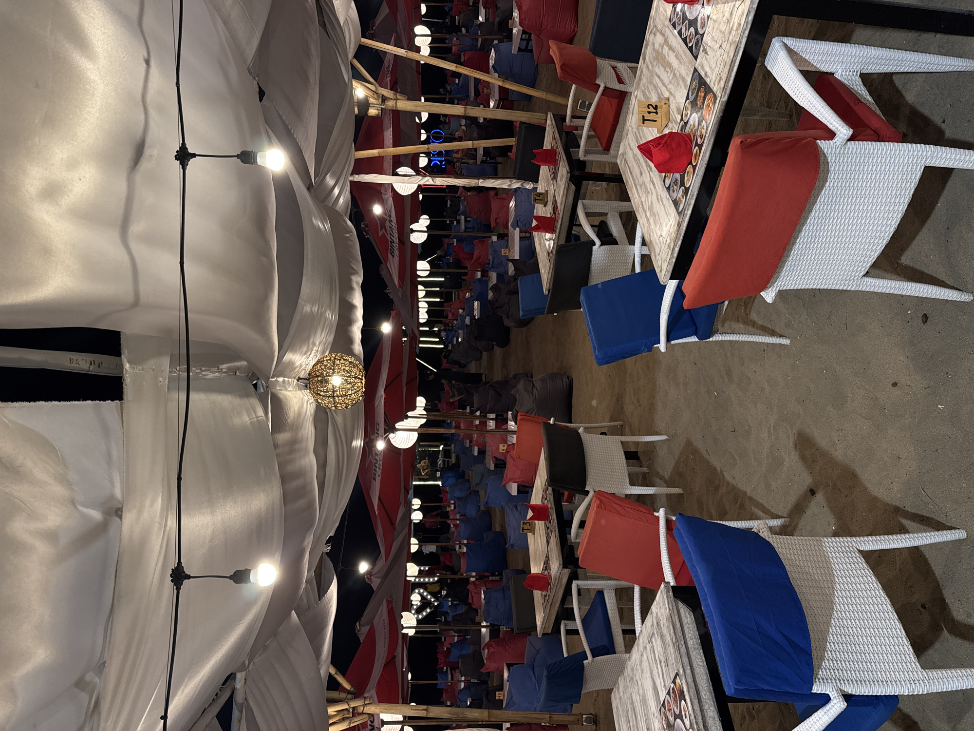 The image depicts an outdoor seating area on a sandy beach, likely in Kuta, Indonesia, during the evening. Numerous tables and chairs, many with colorful cushions (red and blue), are arranged under a canopy of white fabric draped from bamboo poles. Red and white umbrellas are also visible, some branded with "Bintang Crystal." Several beanbag chairs are scattered throughout the area. Lighting is provided by hanging bulbs and a decorative woven orb lamp. The sand is illuminated by the artificial lights, casting shadows from the furniture. In the background, faint figures of people can be seen, suggesting the establishment is operational. The overall scene conveys a relaxed, beachfront dining or lounge atmosphere.