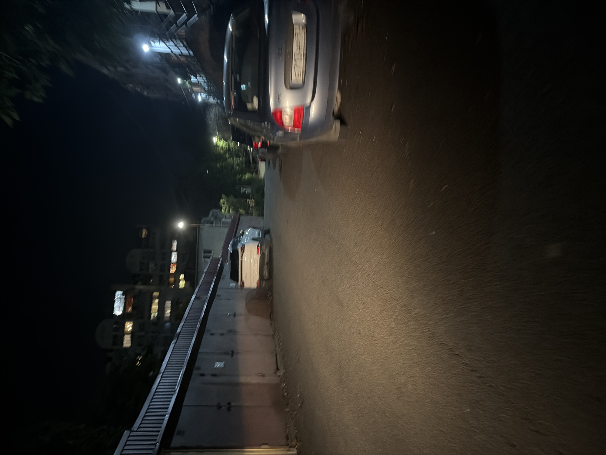 This night-time image, taken in Sochi, Russia, depicts a quiet urban street or pathway. The scene is dominated by artificial lighting from streetlights and building windows against a dark sky.
A light blue or grey hatchback car is prominently parked on the left side of the paved road, with its rear facing the viewer. The car's license plate is clearly visible, displaying the alphanumeric sequence "B712OB" and the Russian regional code "123RUS," which corresponds to Krasnodar Krai, where Sochi is located.
The road surface, likely tarmac, stretches into the distance, appearing somewhat reflective under the ambient light. Further down the path, another lighter-colored car, possibly white, is partially visible.
To the right of the main parked car, several multi-story buildings are dimly visible through the darkness, their various windows illuminated with warm and cool lights. Some dark foliage from trees or bushes partially obscures parts of these buildings and several bright streetlights.
Along the left edge of the road, a metallic railing or fence-like structure runs parallel to the path, suggesting a separation from a lower area or another part of a building complex. On the immediate right side of the road, a textured, light-colored wall or embankment rises, fading into darkness higher up.
The image captures a static moment, with no discernible activity or people, presenting a calm, typical urban night scene.