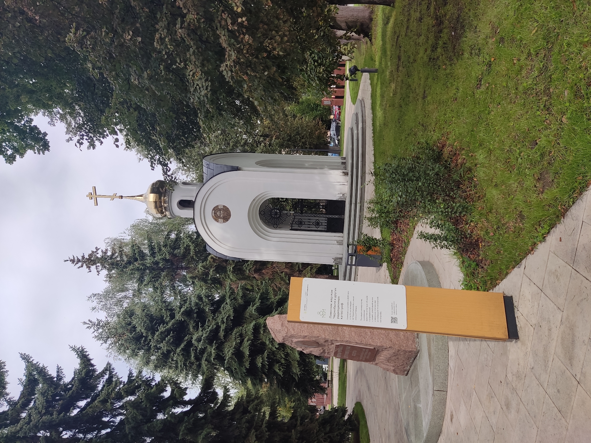 This image depicts a memorial to the victims of Chernobyl and other radiation disasters, located in Noginsk, Russia. The memorial complex features a white chapel with a golden dome and a cross. In front of the chapel, to the left, stands a large stone monument. A wooden information stand with Russian text and a QR code is placed in the foreground, to the right of the stone monument. The surroundings include well-maintained green lawns, trees, and a paved pathway. The sky is overcast. No people are visible in the scene.
