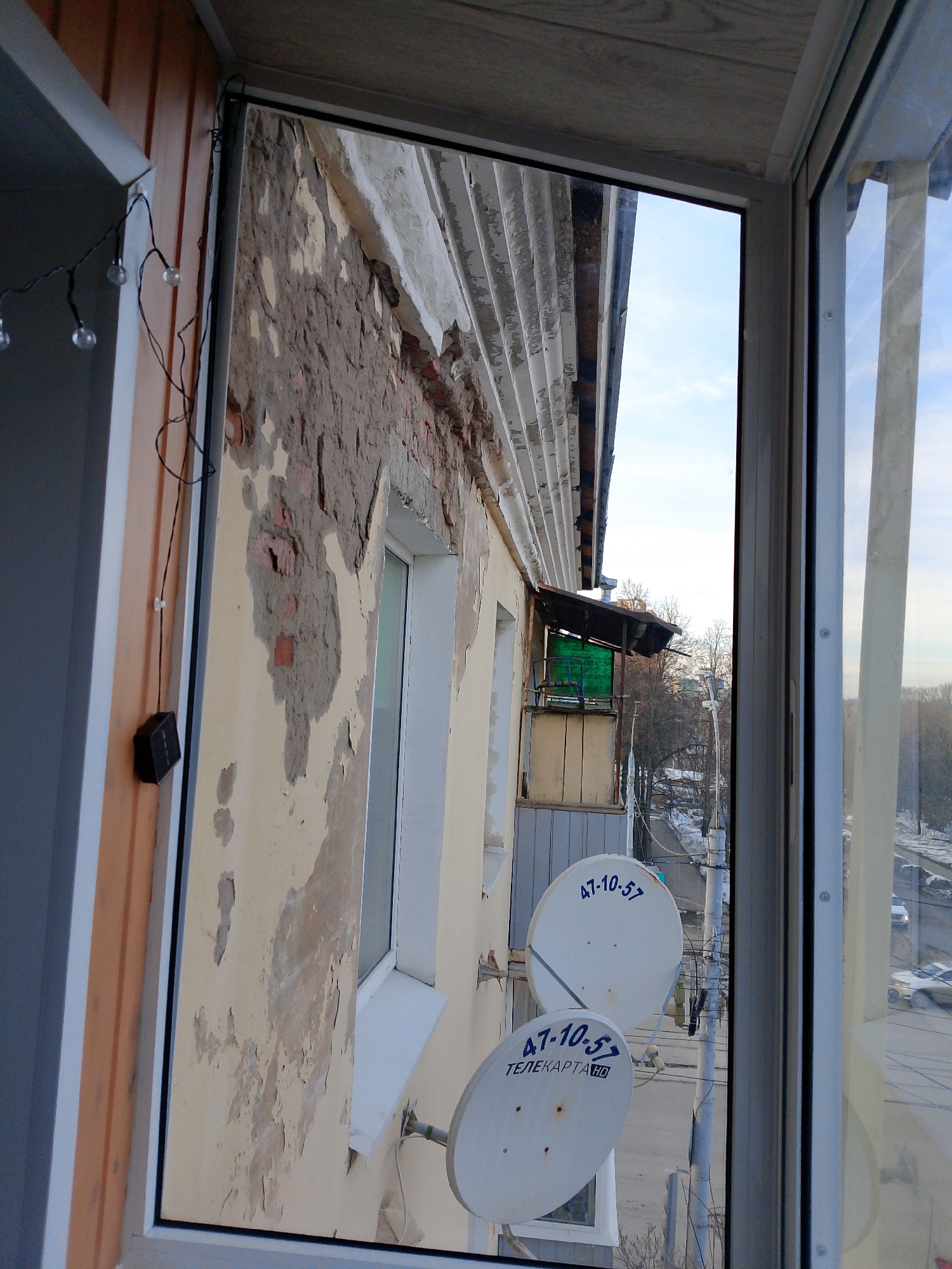 The scene is viewed from an interior space, likely a room or balcony, looking outwards through a white PVC window frame. On the immediate left, an interior wall is clad with vertical orange-brown wooden paneling, from which a string of spherical decorative lights with a black cable hangs. A small black rectangular device, possibly a solar panel, is mounted on this wall below the lights.
The primary external focus is the wall of an adjacent building, which exhibits significant weathering. Its pale yellow or beige paint is extensively peeling and flaking, exposing the underlying grey concrete and patches of red brickwork. A white corrugated metal overhang or roof structure runs along the top of this building. Two white-framed windows are visible on this wall. Below the upper window, two white satellite dishes are mounted. Both dishes display the blue inscription "47-10-57," and the lower dish additionally reads "ТЕЛЕКАРТА HD."
Further in the distance, a darker-roofed balcony with a prominent green side panel is visible. The view extends to a street below, partially covered with snow or slush, lined with bare trees and utility poles carrying electrical wires. Several vehicles are parked along the street. The sky above is a light blue with scattered white clouds. The described location is the city of Izhevsk, Russia.