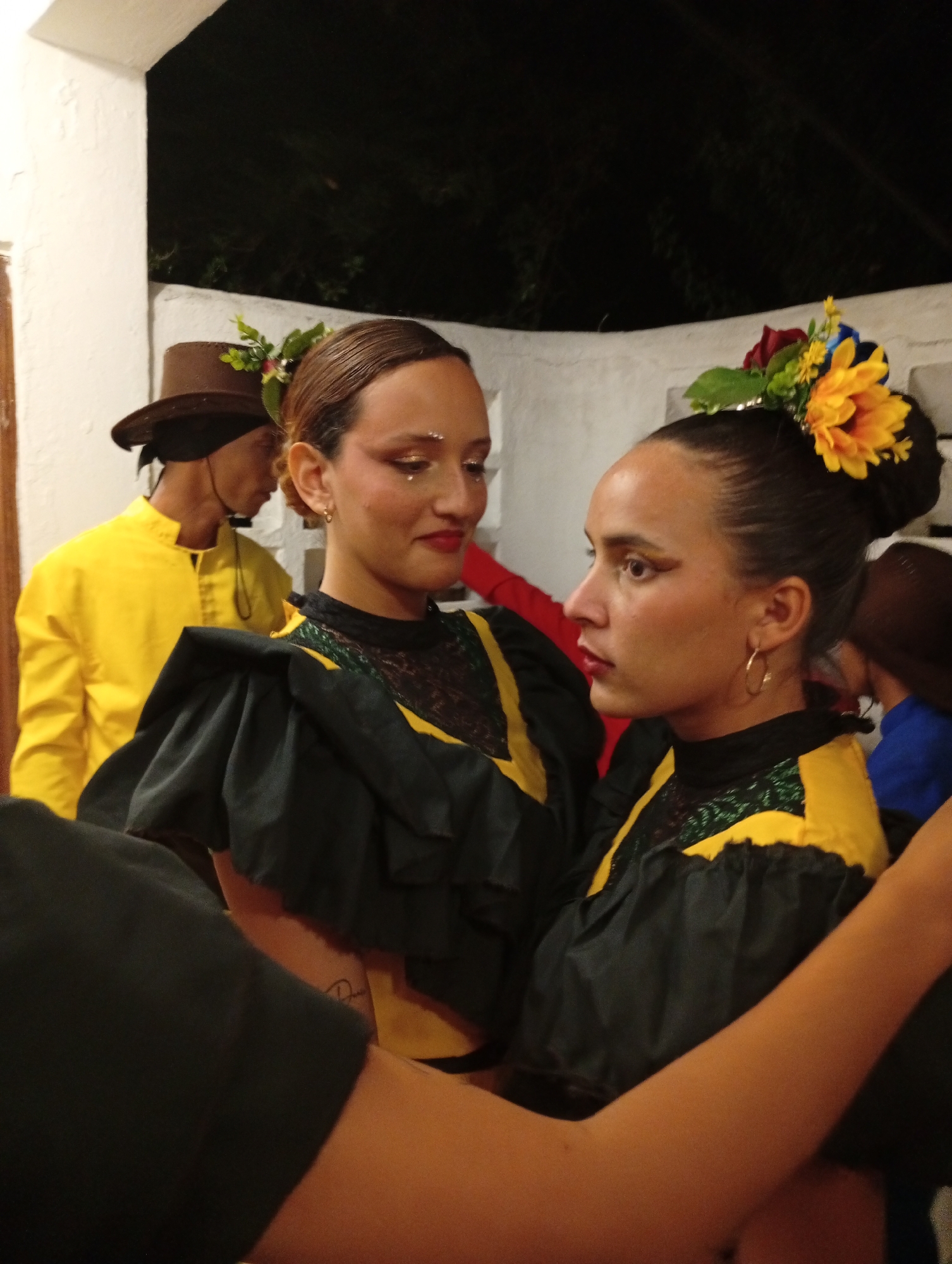 Two women are standing close together in a dimly lit space. Both are wearing black and yellow dresses with ruffled shoulders and floral headpieces. The woman on the right has a large yellow flower in her hair and is looking to the left at the other woman, who is looking back at her. The woman on the left has small, shimmering decorations under her eyes. Both have dark lipstick and defined eyebrows.
Behind them and to the left, a man wearing a yellow shirt and brown hat is visible in the background. A partial view of the interior space is visible including what appears to be a white wall or column and glimpses of other people in the background.
Given the prompt, it can be inferred that the location is Pedro González, Venezuela.