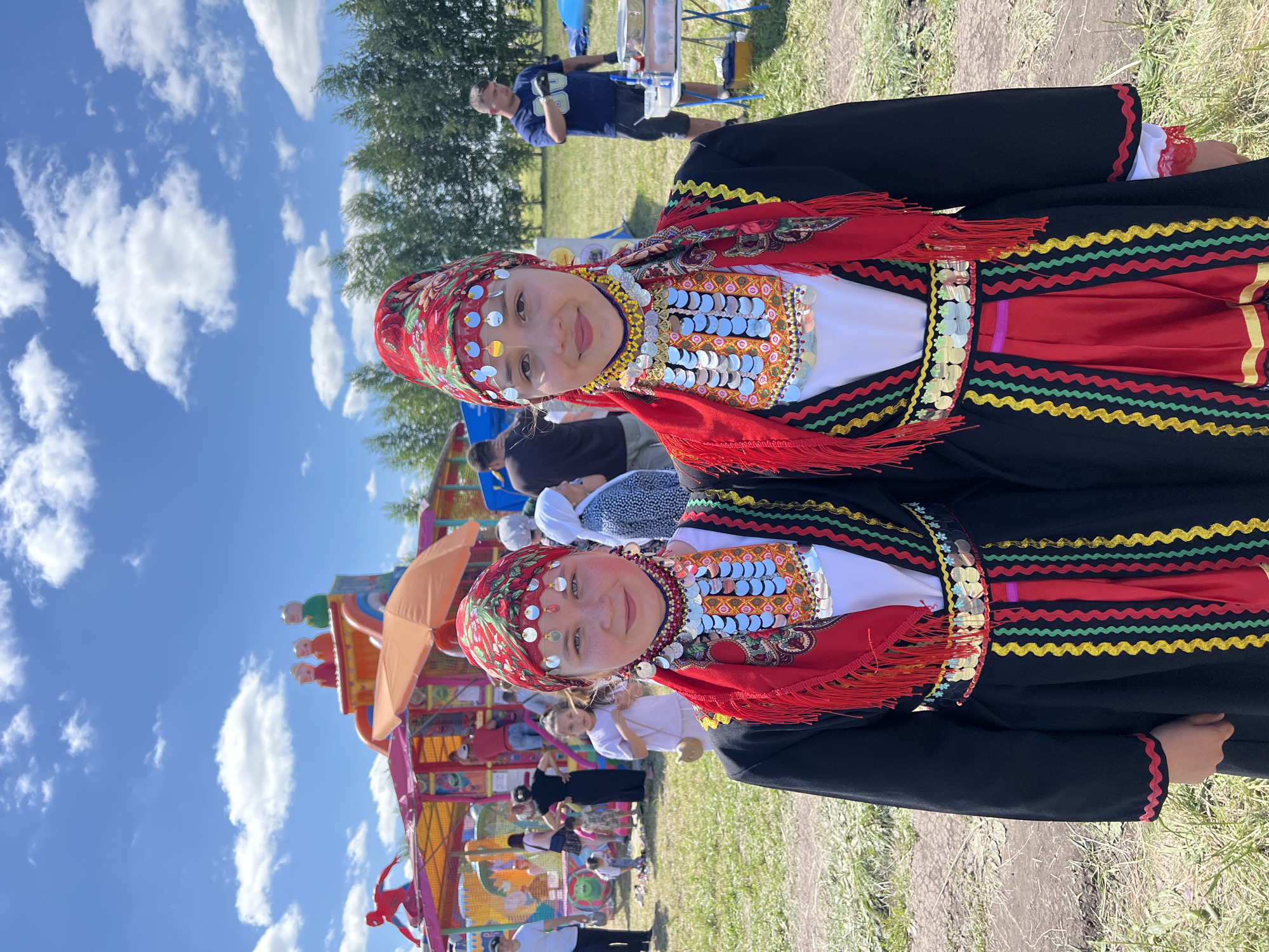 The image depicts a sunny outdoor scene in Staraya Kosa, Russia, likely at a festival or fair. Two young girls, dressed in traditional, brightly colored folk costumes, are the primary subjects, standing close together and looking at the camera. Their attire features red headscarves with intricate patterns, layered necklaces adorned with numerous silver coins and colorful beads, and black tunics over red underskirts with decorative trim. In the background, a lively atmosphere is evident with a large, colorful inflatable playground structure, a crowd of people of various ages milling about, and a table set up with food or supplies. The overall setting suggests a community gathering on a clear, blue-sky day.