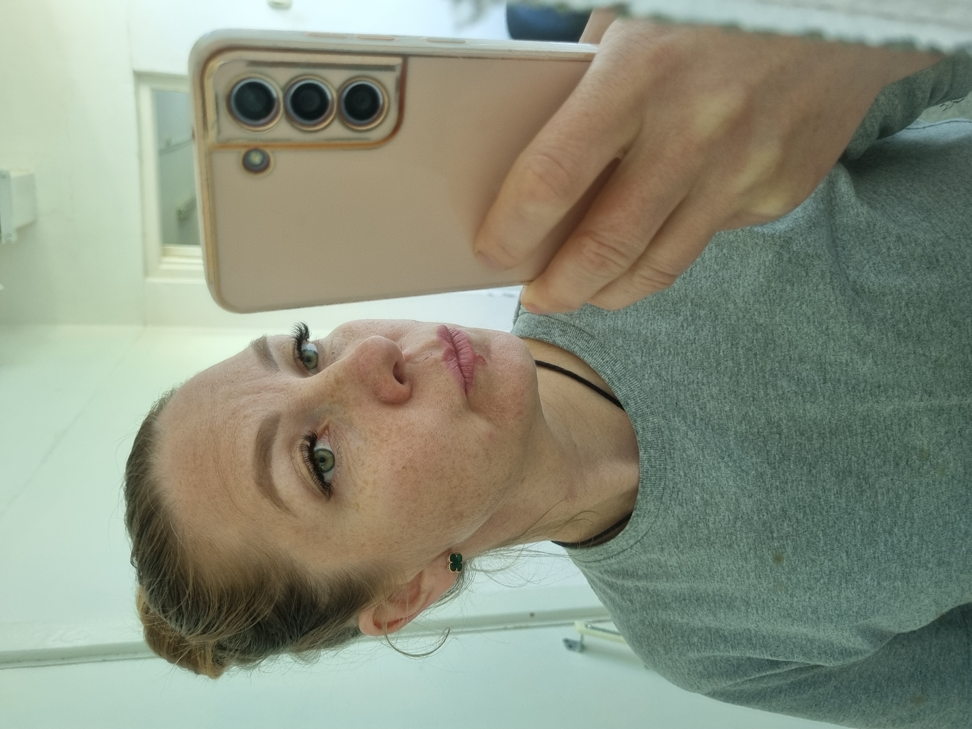 A woman is taking a selfie with a pink smartphone. The phone has three rear cameras. The woman has freckles, green eyes, and light brown hair pulled back in a bun. She is wearing a grey long-sleeved shirt and small, clover-shaped earrings. The background appears to be a white or light-colored room with a window. There is no information to suggest the location is in Kemul, Russia.