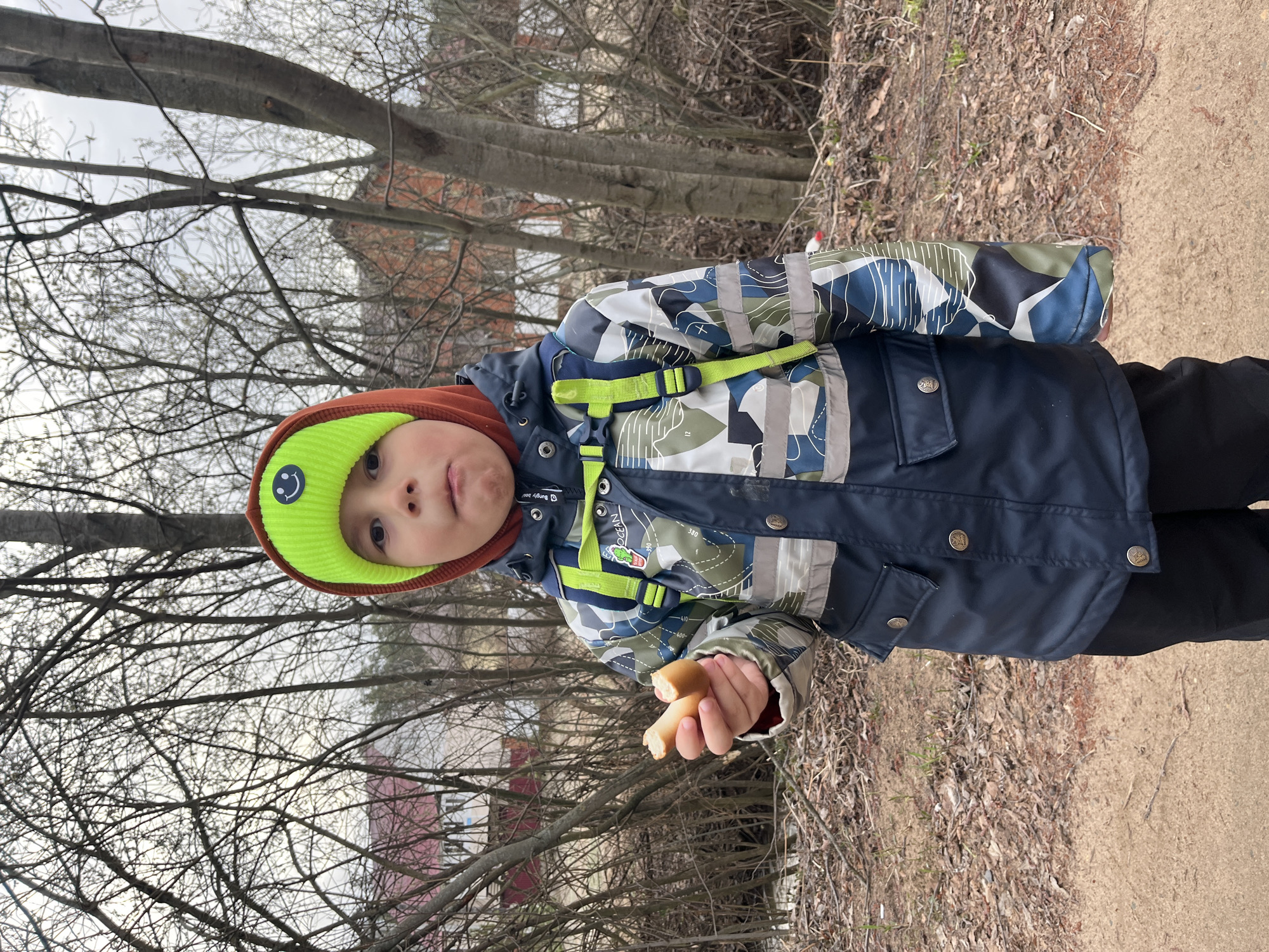 A young child stands outdoors, holding a piece of bread or similar food item in their right hand. The child wears a brown and bright green beanie with a smiley face emblem, and a camouflage jacket with a dark blue lower portion. Behind the child are bare trees and what appears to be a building with red elements in the background. The ground shows signs of autumn, with dead leaves and bare soil. The image was taken in Chaikovsky, Russia.