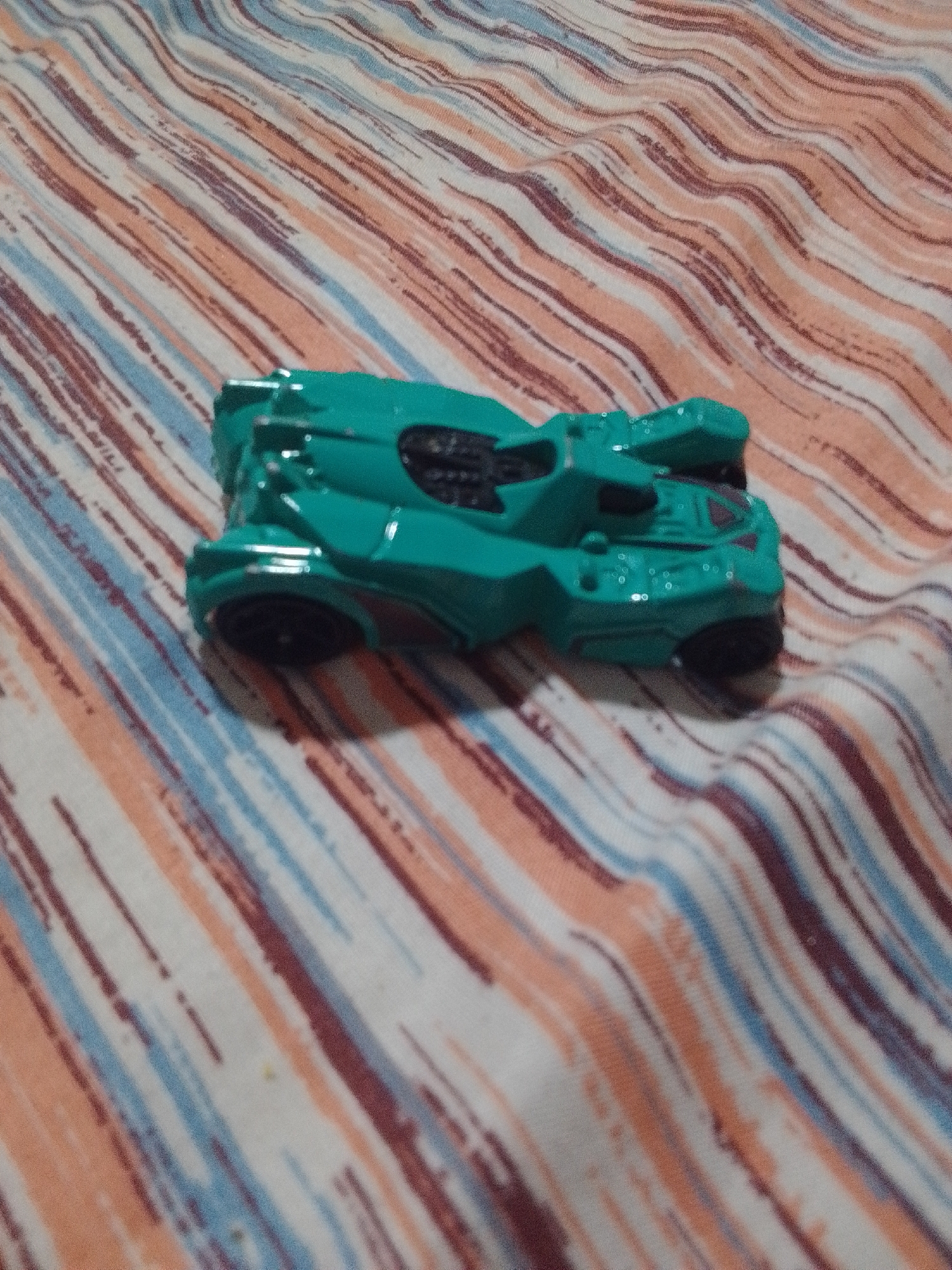 A teal toy car is positioned diagonally on a striped fabric, likely a blanket or bedsheet. The fabric features diagonal stripes of white, light orange, and dark blue, with some areas appearing slightly darker or lighter due to the fabric's texture and folds. The toy car is a stylized vehicle, possibly inspired by a superhero car or a futuristic concept. It has a sleek, angular design with visible wheels and body details. The car is the primary subject, with the fabric serving as the background and setting. The lighting suggests an indoor environment, possibly during the daytime, but no specific time of day or weather conditions can be determined. There is no visible text in the image. The overall impression is a simple, static scene of a toy on a textured surface.