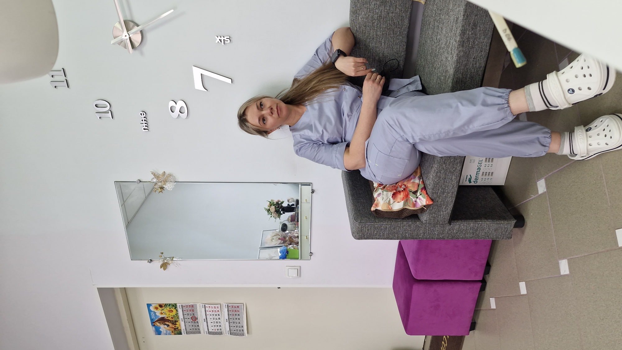 An indoor full-shot depicts a woman in light blue medical scrubs and white Crocs, sitting casually on a gray two-seater couch. She has long blonde hair and is looking directly at the camera with a neutral expression. Her left arm rests on the armrest, while her right hand is clasped over her left knee. A floral-patterned cushion provides back support.
The setting appears to be a professional space, likely a beauty salon, dermatology clinic, or waiting area in Otchor, Russia. The walls are light gray. Behind the woman, a large, modern wall clock features metallic numbers "11," "10," "8," "7," and the words "nine" and "six." The clock hands indicate approximately 10:15. Below the clock, a rectangular mirror, adorned with decorative golden accents, reflects parts of the room including a shelf with various items and floral arrangements. To the left of the mirror, a wall calendar for 2026 is visible, showing "АПРЕЛЬ" (April) and "ИЮНЬ" (June).
Next to the couch, a product box for "dermaGEL" is placed on the floor or a low surface, displaying pH level numbers such as "4.0," "5.0," "6.0," "7.0," "8.0," "8.5," and "9.0." In the lower right of the frame, parts of purple upholstered seats or ottomans are visible. A white pen or tool with the number "909" is partially visible near her feet. The floor is light-colored tile with some visible seams and white square markers.