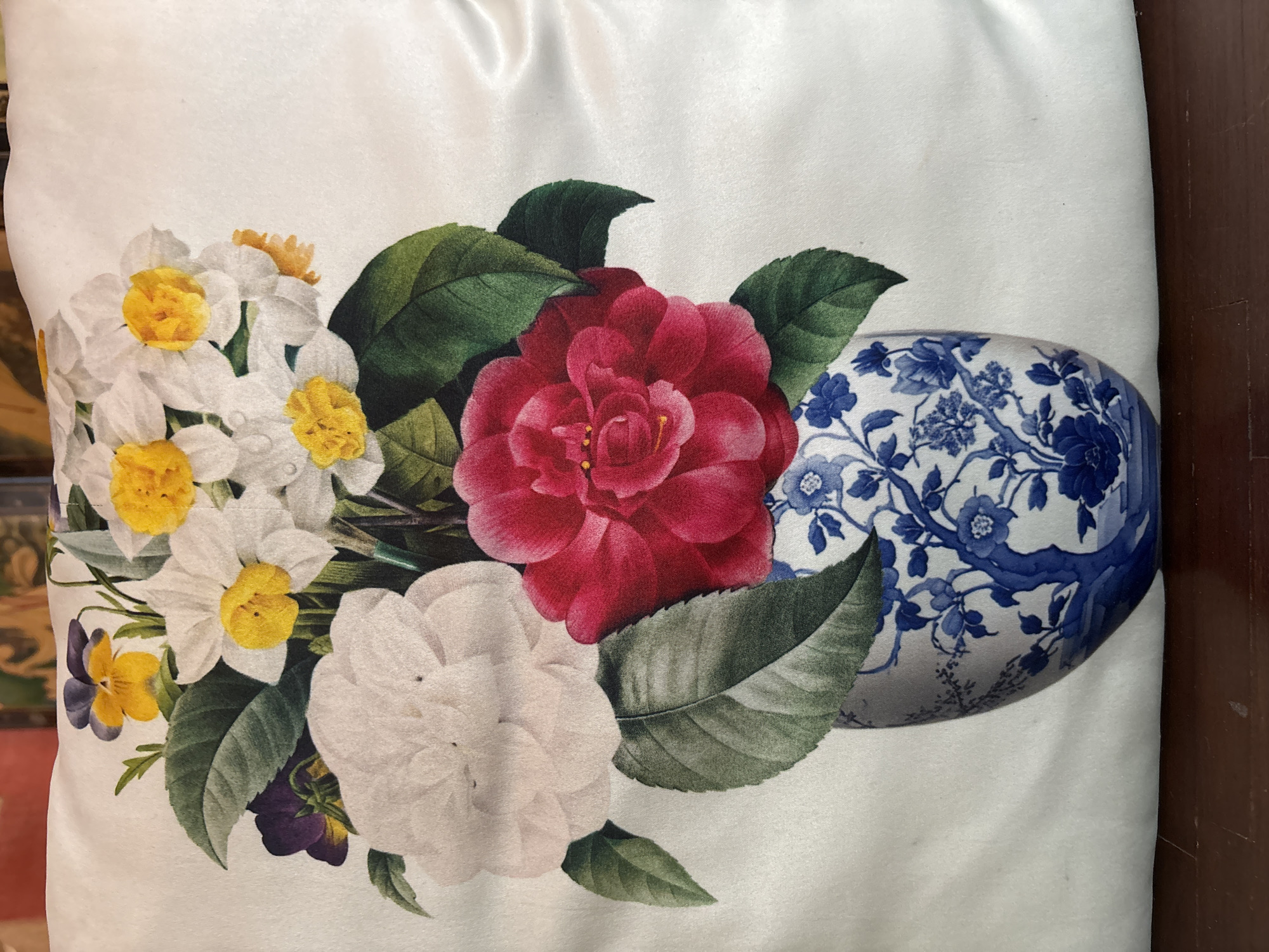 The image is a close-up shot of a decorative pillow with a botanical print. The print features a vibrant arrangement of flowers, including white and pink camellias, yellow daffodils with white petals, and small purple and yellow pansies. These flowers are arranged in a blue and white porcelain vase with a detailed floral and tree pattern, reminiscent of Ming dynasty style ceramics. The background of the pillow is a plain white, which appears to be made of satin fabric, reflecting light and creating a soft sheen. The lighting suggests an indoor setting. There is no indication of the location being in the city of Chalong, Thailand; the image shows an inanimate object.