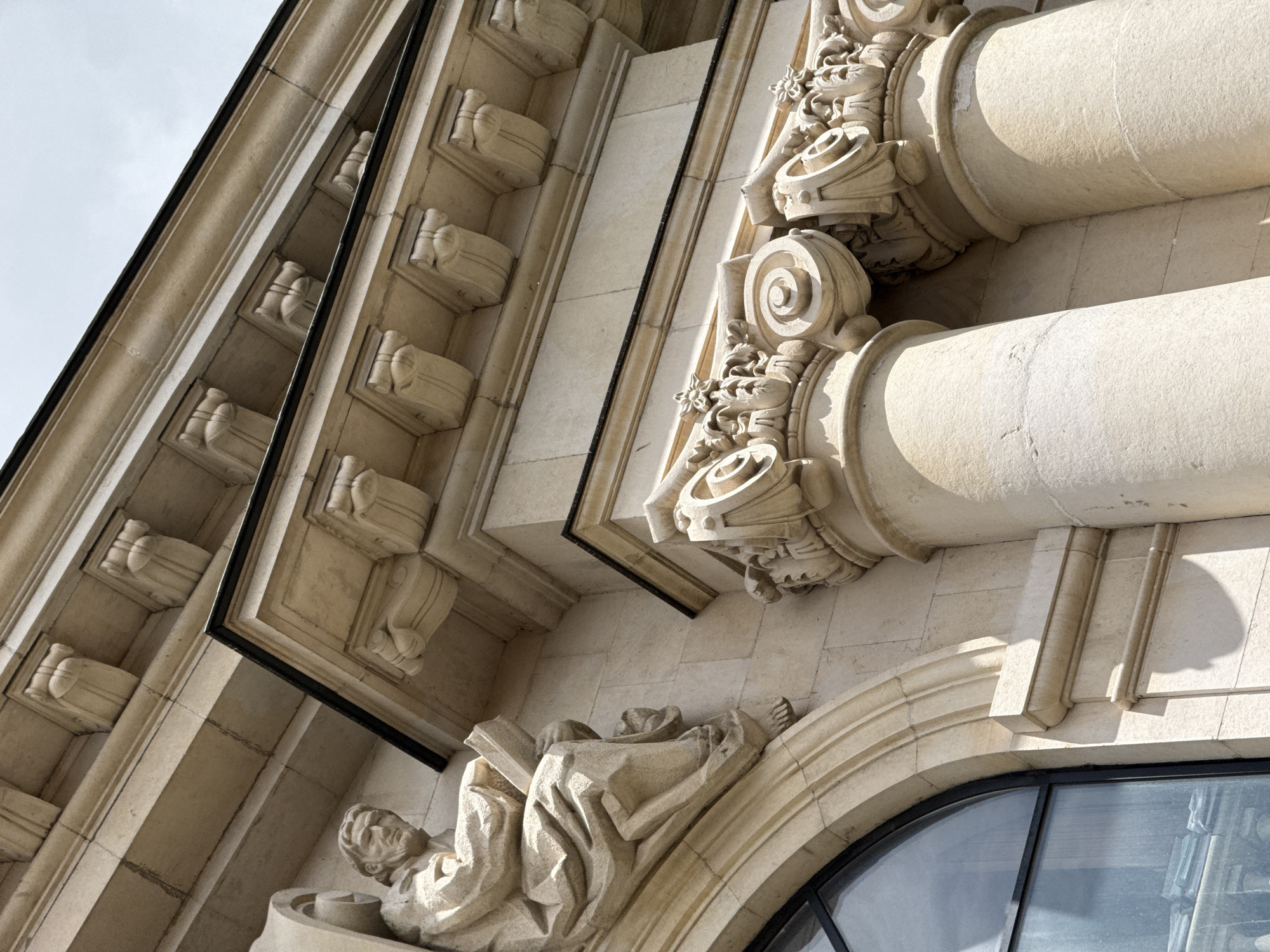 The image shows a close-up view of a building facade, featuring architectural details and sculptures. The building is constructed of a light-colored stone or concrete. The facade is adorned with decorative elements, including columns, molded cornices, and carved ornaments.
A prominent feature is a sculpted figure of a person, positioned above an arched window. The person is reclining and appears to be holding a book.
The photograph is shot from a low angle, looking upwards, capturing the interplay of light and shadow on the building's surface. The sky is partially visible in the background.
Without additional landmarks or clues, it is difficult to confirm the specific location in Sofia, Bulgaria, but the architectural style suggests a historical building.