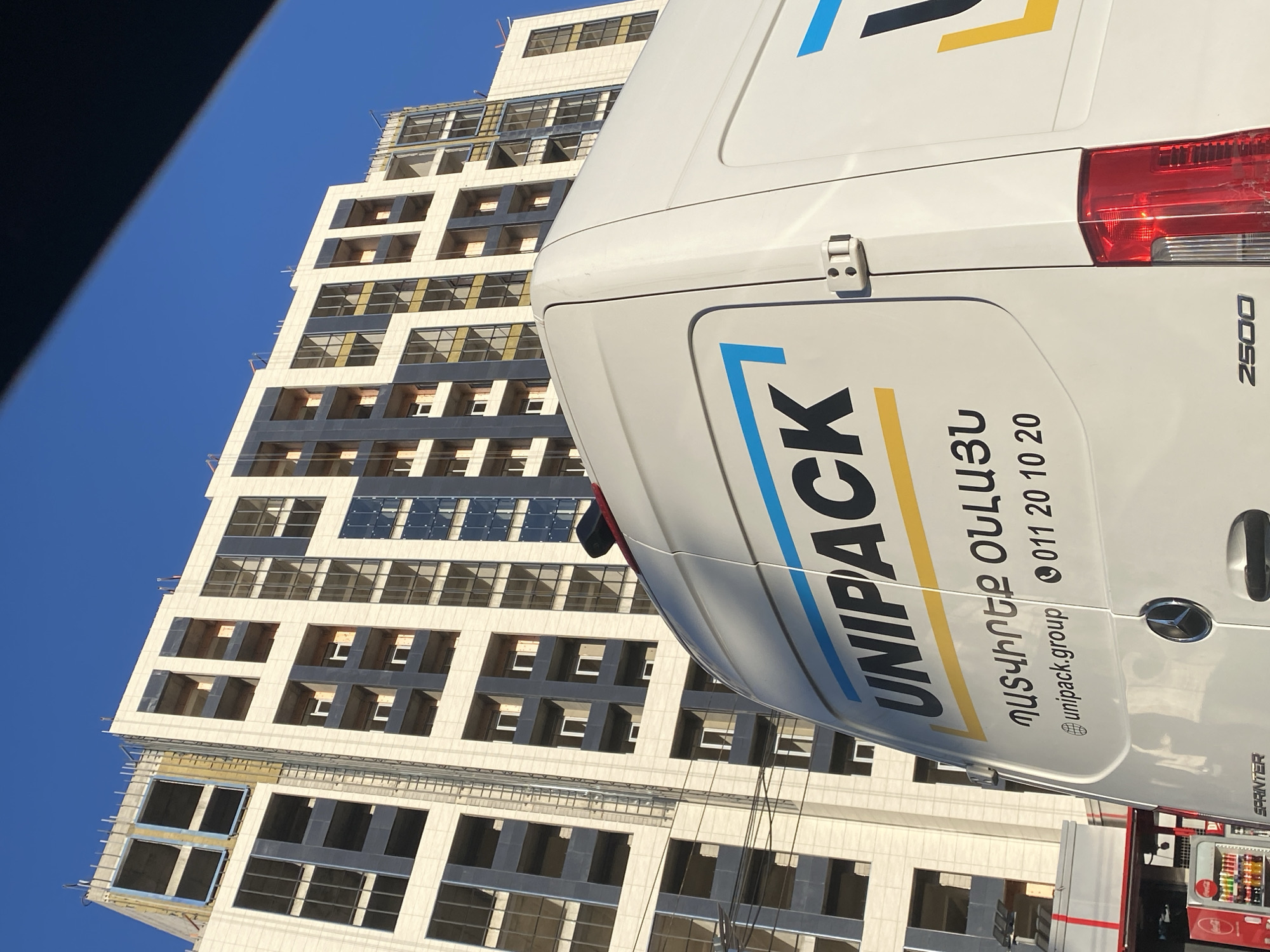 The image shows the side of a white Mercedes-Benz Sprinter van with the text "UNIPACK" and Armenian text printed on it. The van is parked in front of a modern building under construction. The building has a facade of light-colored stone and dark windows, with scaffolding visible. The sky is clear and blue, suggesting a sunny day. The image is taken from a low angle, looking up at the van and the building. The time of day appears to be late morning or early afternoon based on the sunlight. The overall scene depicts an urban environment in Yerevan, Armenia, with a focus on commercial vehicles and ongoing construction. The text on the van provides branding and contact information for the company Unipack.