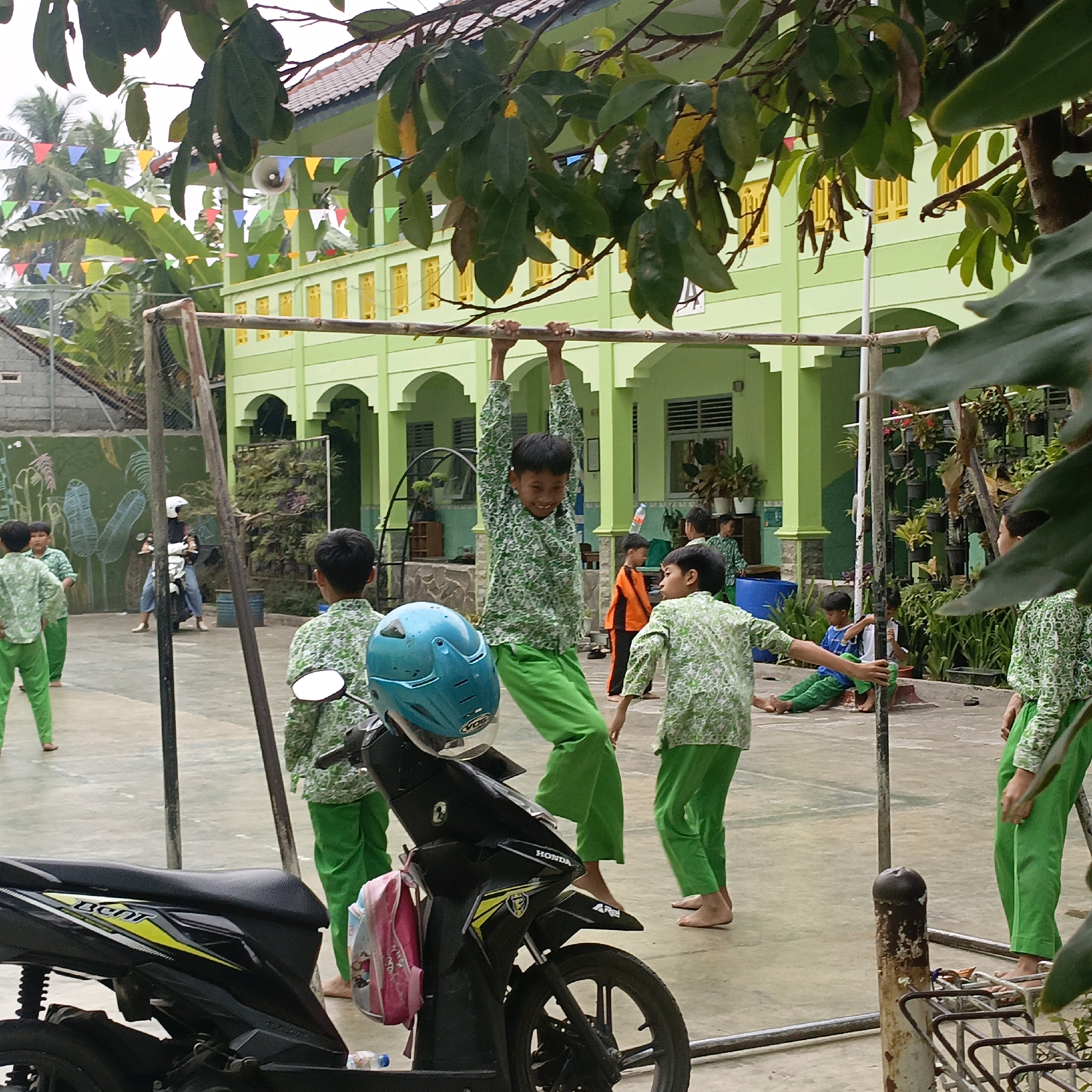 In Salatiga, Indonesia, at a school with a light green building and yellow window frames, a group of young boys in matching green patterned shirts and bright green pants are gathered in an outdoor play area. The area is paved and features a horizontal bar for gymnastics. One boy is actively hanging from the bar, smiling broadly, while others observe. In the foreground, a black motorcycle with a blue helmet on its seat is parked. The scene is framed by the leaves of a large tree. The weather appears overcast.