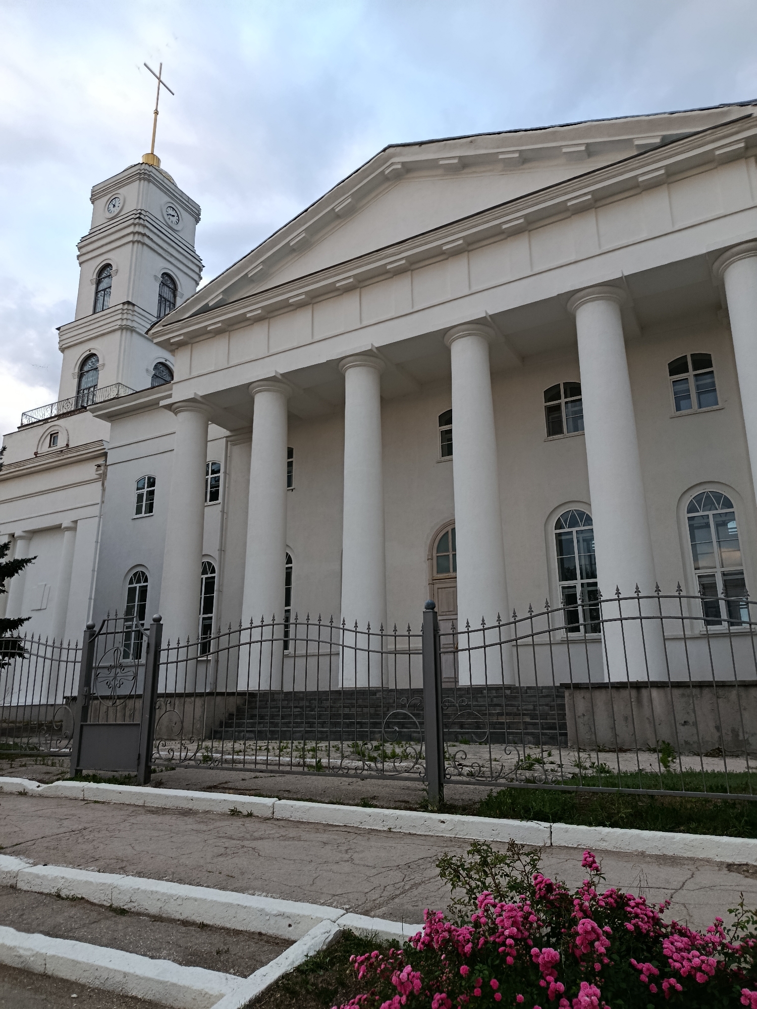 The image shows the exterior of a white classical-style building with a prominent portico supported by large columns. A tall, white bell tower with clock faces is visible on the left side of the building, topped with a golden cross. A metal fence runs along the front of the property, with a gateway in the foreground. Lush green grass and some pink flowering bushes are present in front of the fence. The sky is overcast. The building is identified as the Holy Ascension Cathedral, located in the city of Marx, Russia. There are no people or animals visible in the image.