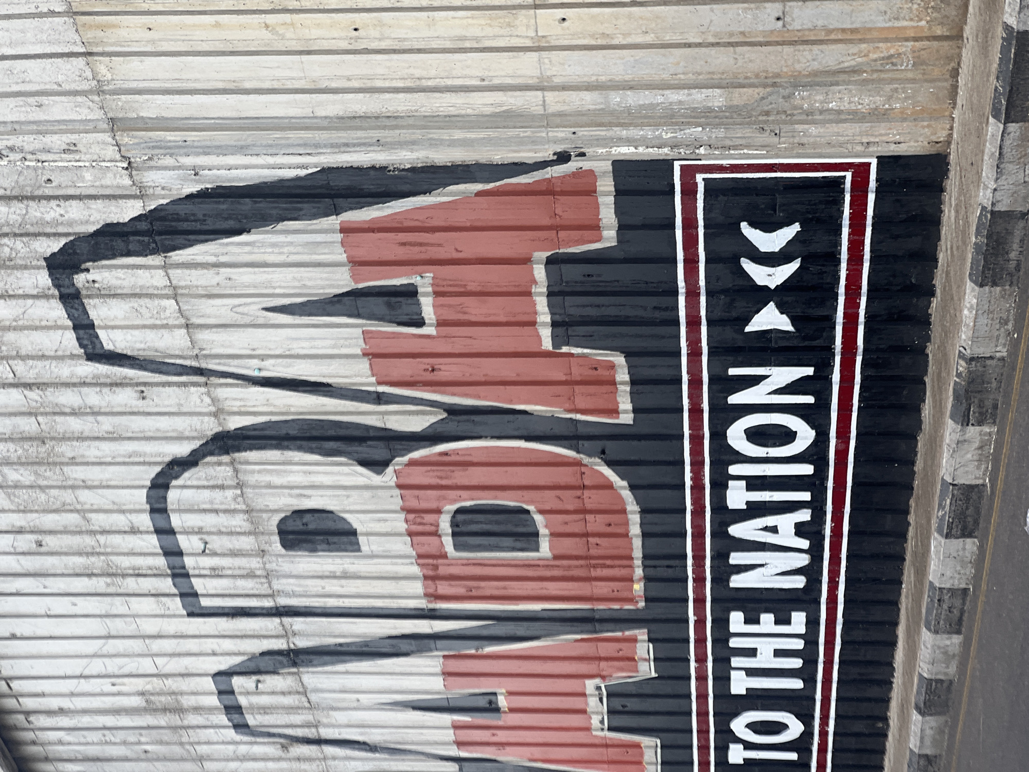 This image shows a mural painted on a corrugated metal wall. The mural prominently features the word "ABA" in large, bold letters, with each letter filled with red and outlined in black. Below "ABA," there is a horizontal banner with white lettering that reads "TO THE NATION," bordered by red and black stripes. The wall itself appears to be part of an outdoor structure, possibly a building facade or a large fence, and shows signs of weathering and age. The ground in front of the wall is a paved road with a grey and black checkered curb. This mural is located in Jalingo, Nigeria, serving as a visible piece of public art or branding.