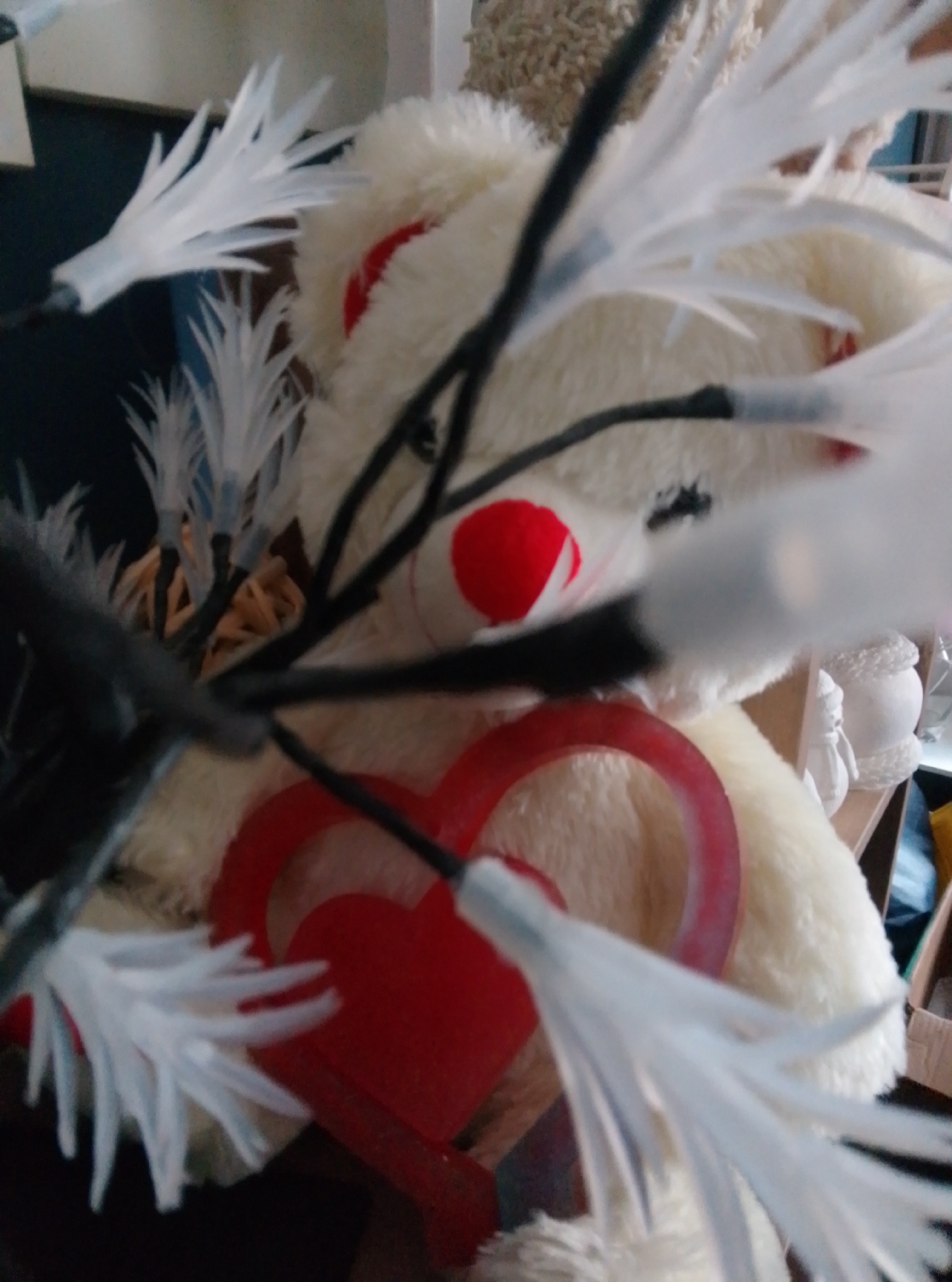 A close-up, medium-shot photograph captures a fluffy, cream-colored teddy bear. The bear is adorned with a prominent bright red heart-shaped decoration around its neck, with a section of a second, larger red heart visible below. The bear's nose is also a bright red sphere. The bear is partially obscured by a cluster of delicate, white, feather-like artificial branches, some of which appear to have small, clear lights attached. These branches are arranged in a way that suggests they are part of a decorative display. In the background, out of focus, are what appear to be shelves holding various objects, including a light-colored ceramic globular item and a cardboard box. The lighting is uneven, with some areas brightly lit and others in shadow. The photograph was taken indoors, likely in a domestic setting, and there is no indication of the city of Wedel, Germany.