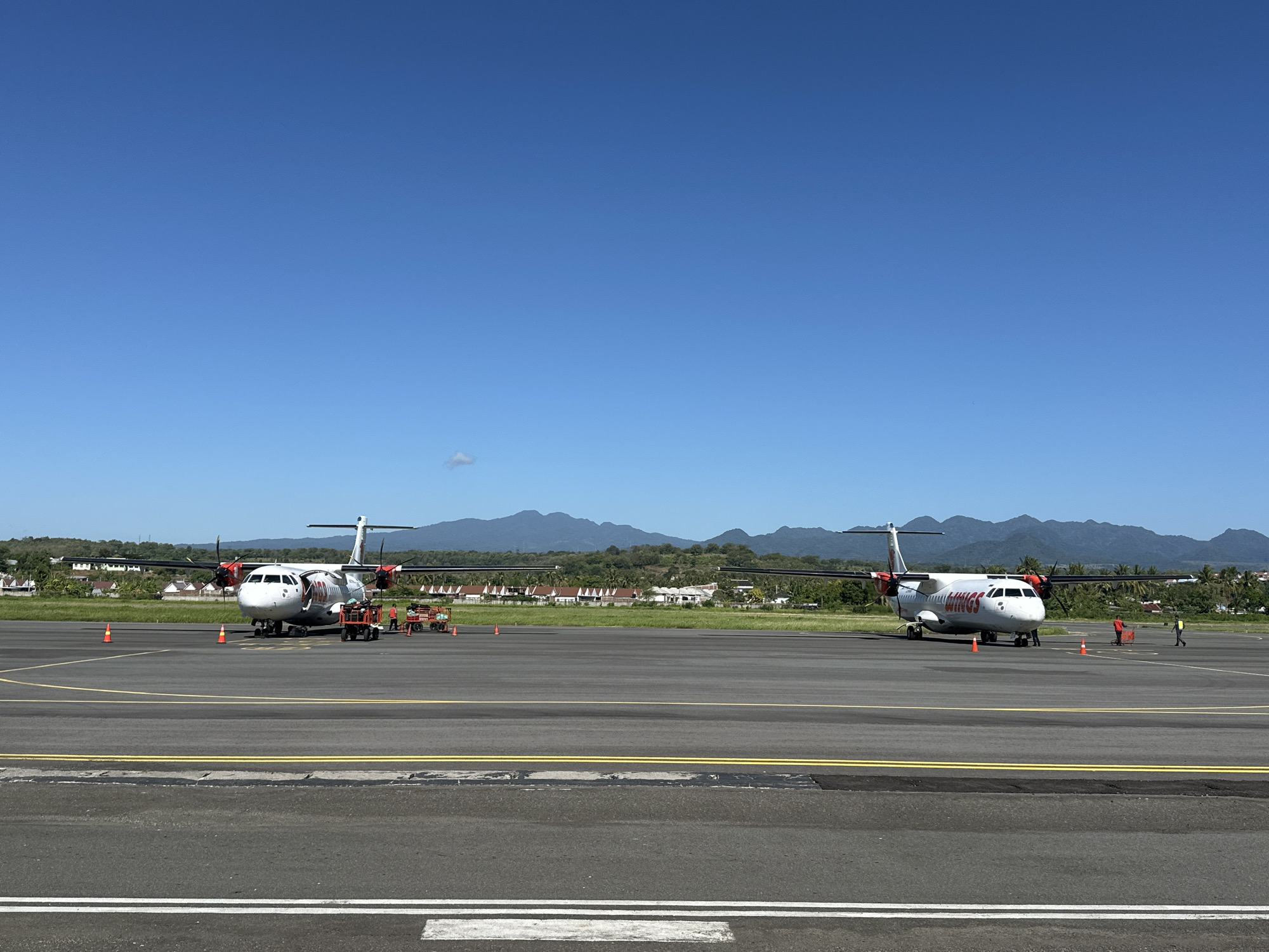 Two turboprop aircraft are parked on an airport tarmac under a clear blue sky. The aircraft on the left is white with red and black accents, and the word "Wings" is visible in red lettering on its fuselage. It is flanked by two orange traffic cones and appears to be undergoing service, with a small utility vehicle and several individuals in orange vests standing nearby. The aircraft on the right is also white with red and black accents, and the word "Wings" is visible in red lettering on its fuselage. It is situated further down the tarmac, and two individuals in orange vests are visible near it. The tarmac is marked with yellow and white lines. In the background, there is a strip of green grass, followed by a line of houses with red roofs and palm trees, and beyond that, a range of rolling, tree-covered hills and mountains. The scene is a typical view of an airport in a tropical or subtropical location. The location is the Lempeh Airport (LMP) in Lempeh, Indonesia, based on the distinctive landscape and vegetation.