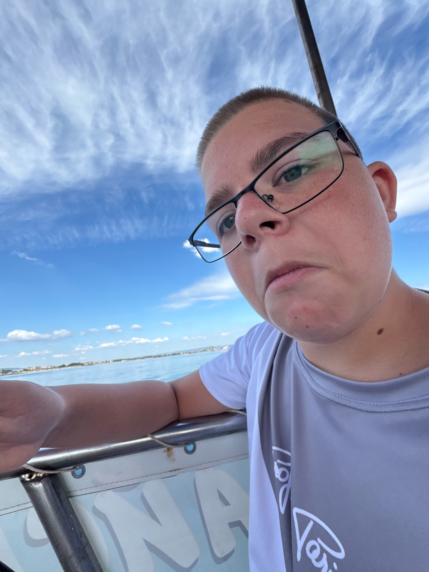 A close-up selfie shows a person wearing glasses and a grey t-shirt, leaning on a railing. The person has short hair and appears to be on a boat, with the sea and a distant coastline visible under a cloudy blue sky. The visible part of the coastline suggests the person is in or near Nesebar, Bulgaria, a city known for its historic old town on a peninsula. The railing has some rope tied to it, and a partial logo is visible on the side of the boat.