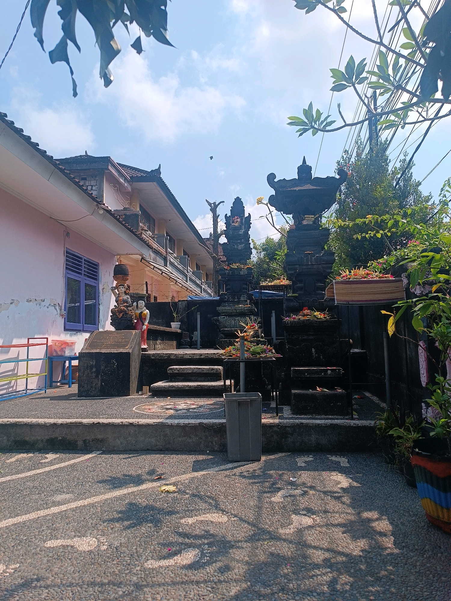 The image depicts an outdoor scene in Tabanan, Indonesia, with a Balinese-style temple structure as the central focus. The temple features dark stone carvings, tiered platforms, and offerings of flowers and incense. To the left, a light pink building with purple-trimmed windows is visible, suggesting it could be part of a school or residential complex. In front of the building, a dark gray plaque is mounted on a stone base, flanked by two statues of women carrying items on their heads. Steps lead up to the temple area, which is adorned with colorful floral offerings. The ground is paved with small stones, and shadows from overhead foliage are cast across it. Trees and lush greenery surround the temple, and power lines are visible overhead. The sky is blue with scattered clouds, indicating a sunny day. A gray trash can is positioned to the left of the temple steps.