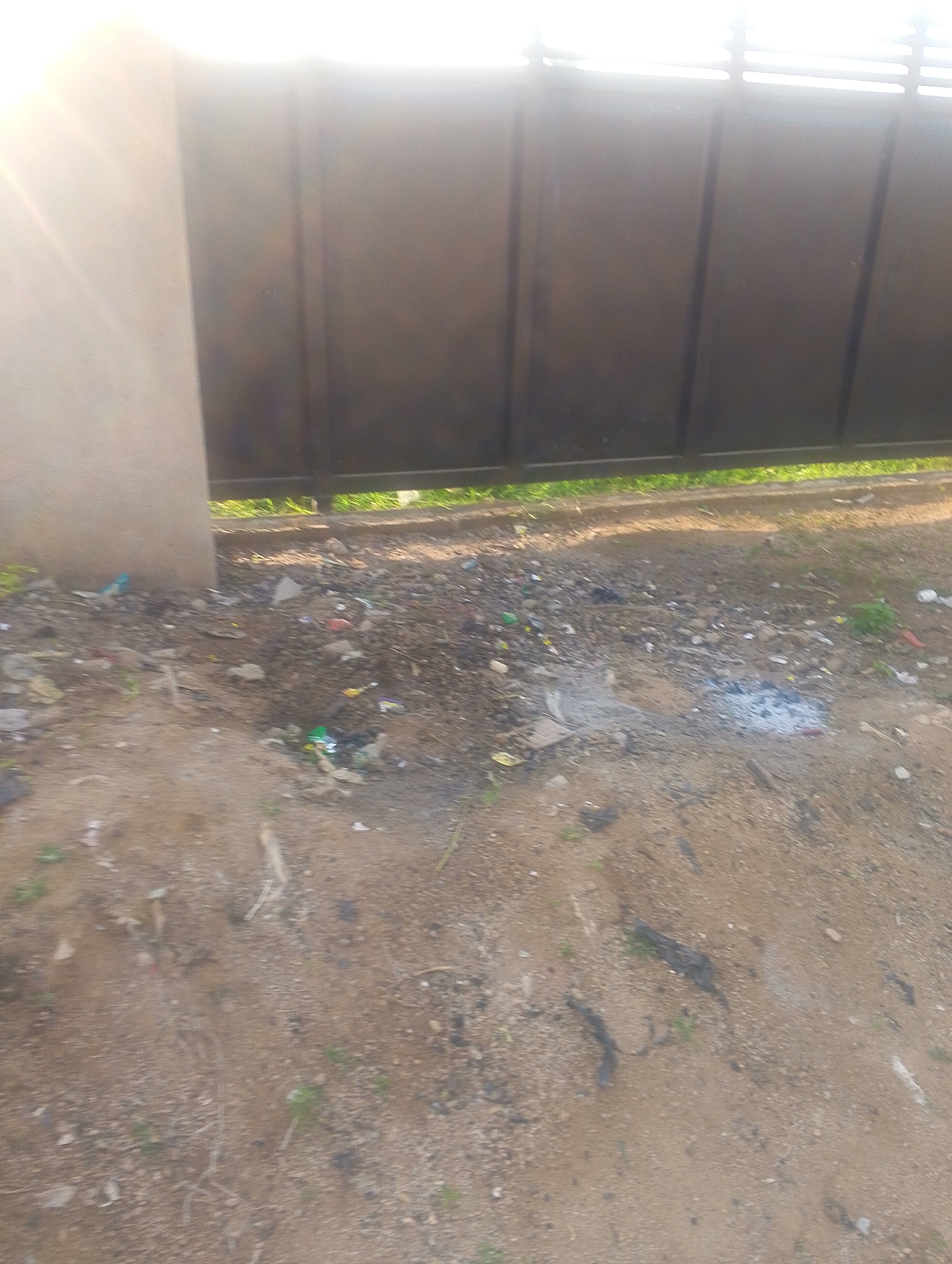 The image depicts an outdoor scene in Jalingo, Nigeria, featuring a large, dark metal gate with horizontal slats at the top. To the left of the gate is a light-colored, textured pillar. The ground in front of the gate is covered with soil and debris, including small pieces of trash such as plastic wrappers and what appear to be cigarette butts. There is also some sparse green vegetation growing among the debris. The lighting suggests it is daytime, with bright sunlight creating a glare on the left side of the image and casting shadows. The overall environment appears unkempt and suggests an area where waste has accumulated.