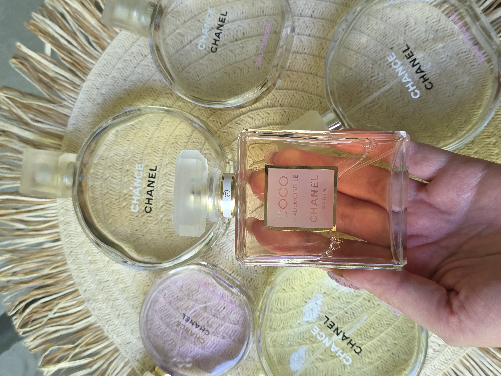 This is a top-down, eye-level shot featuring several bottles of Chanel perfume arranged on a textured, woven surface with a fringed edge. The focus is on a hand holding one of the perfume bottles, a rectangular glass bottle labeled "COCO MADEMOISELLE CHANEL PARIS". The other visible bottles are round and appear to be part of the "CHANCE" line by Chanel, with labels indicating "CHANCE EAU TENDRE" on one, and "CHANCE CHANEL" on others. The perfumes vary in color, with one appearing pinkish, another pale yellow, and two others with a pale pinkish hue. The lighting is bright and natural, suggesting daylight. The overall aesthetic is luxurious and elegant, with a focus on the Chanel brand. The hand holding the "COCO MADEMOISELLE" bottle is lightly colored with manicured nails, adding to the refined presentation. The background is softly blurred, keeping the attention on the perfumes and the hand. There are no clear indications of the specific time of day, weather, or geographical location, but the scene evokes a sense of personal grooming or a beauty product display.