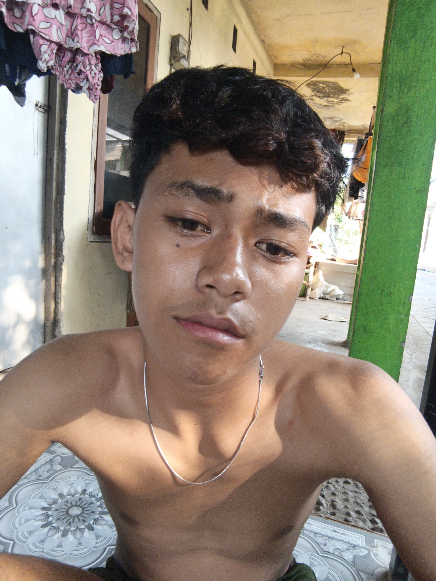 A young man with dark, curly hair, a silver necklace, and a faint mustache is shown from the chest up, facing the camera. He has clear skin, a mole near his right eye, and is shirtless, exposing his shoulders and upper chest. He is outdoors, with a patterned tiled floor beneath him and a green post to his right. In the background, there are clothes hanging on a line, a window with floral curtains, and a yellow-painted wall and ceiling. The scene appears to be in an outdoor living area or porch.