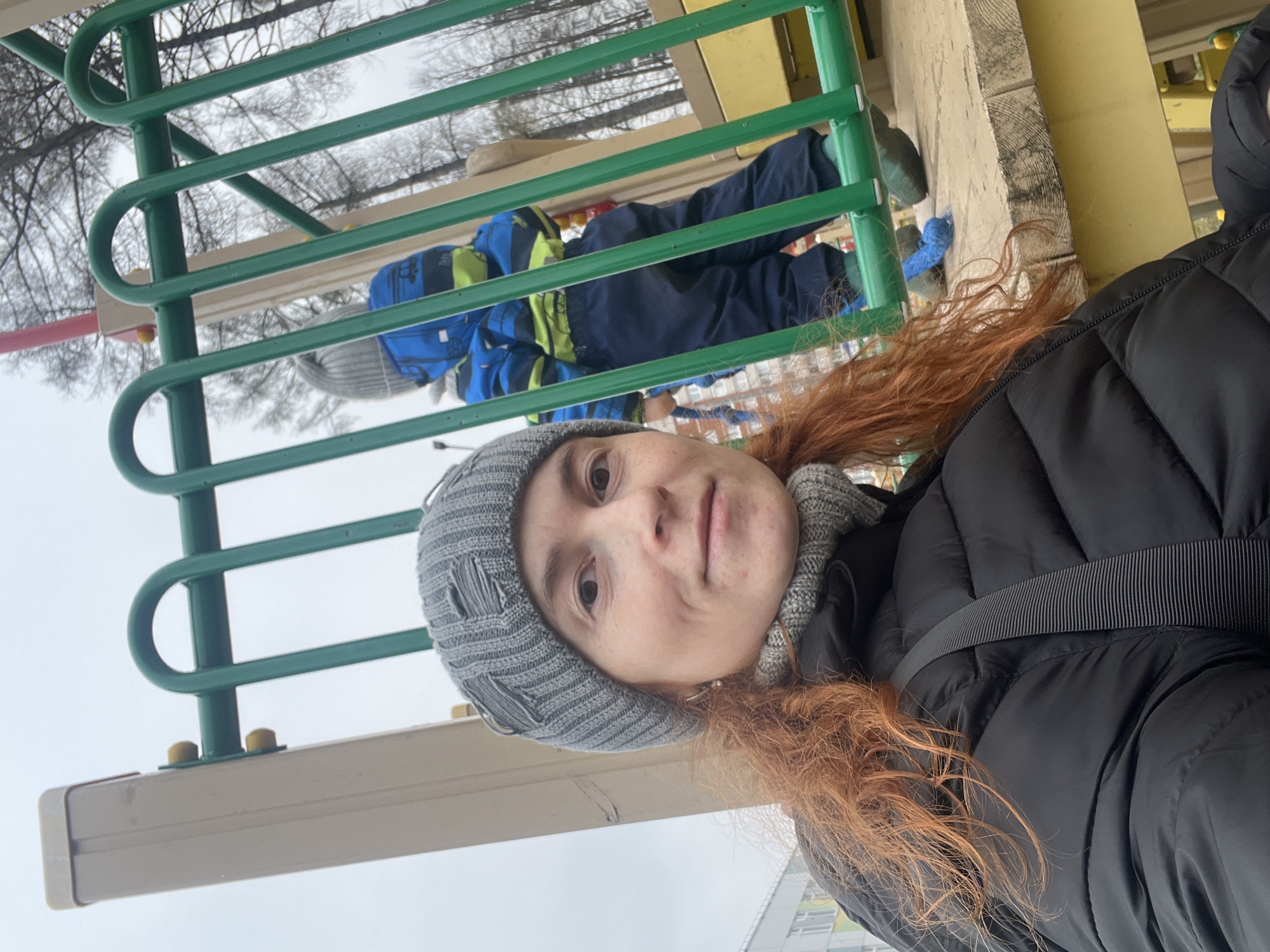 A selfie captures a woman at an outdoor playground in Chaikovsky, Russia. The image is oriented vertically.
**Subjects:** The primary subject is a woman in the foreground, with reddish-brown, curly hair flowing around her shoulders. She wears a grey knitted beanie with distressed elements, a matching grey knitted scarf, and a black puffer jacket with a textured black strap across the mid-section. She looks directly at the camera with a neutral to slightly smiling expression. In the background, partially obscured by green metal playground equipment, is a young child. The child wears a blue winter jacket with bright neon yellow accents, blue pants, and a grey knitted hat, and appears to be standing on the playground structure.
**Scene and Setting:** The setting is an outdoor playground, characterized by the prominent green metal bars of a climbing frame and a light-colored wooden or composite platform. The background reveals bare trees against a pale, overcast sky, indicative of a cold season like winter or early spring.
**Activity:** The woman is taking a selfie, likely while supervising or accompanying the child who is engaged with the playground equipment.
**Notable Details:** The weather appears cold and overcast, necessitating warm winter attire for both individuals. The trees in the distance are bare, reinforcing the seasonal context. No discernible text is visible on any subject or object within the frame.