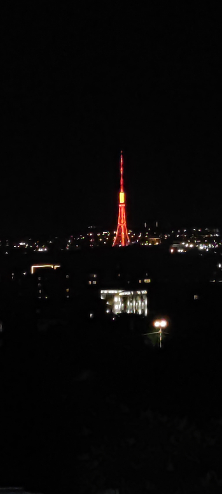 The image presents an elevated, nighttime view of an urban landscape, identified as the city of Yerevan, Armenia. Dominating the scene is a tall, slender lattice tower, brightly illuminated in red and orange, with a more intensely lit yellow-orange section at its upper portion. The tower's intricate structure is clearly delineated by the lights against the dark, clear sky. Below the tower and extending across the mid-ground, the city unfolds with numerous scattered points of light from buildings and unseen thoroughfares, indicating active urban life. A distinct, brightly lit building with multiple windows or vertical elements is visible in the mid-ground, slightly to the right of the tower's base. Further in the foreground on the right, a streetlamp with two prominent, round yellow lights provides additional illumination. The overall environment is dark, with artificial lights defining the visible elements. No human figures are present, and no specific actions are discernible.