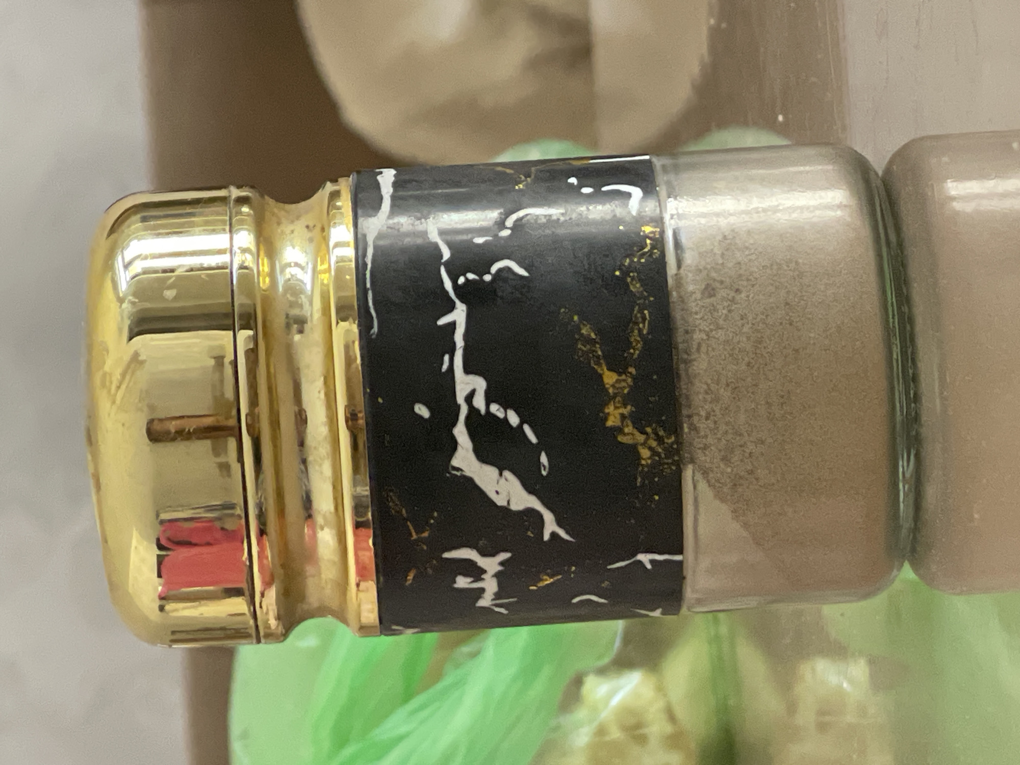A close-up shot focuses on a small glass jar filled with a light brown powder, possibly spices or makeup. The jar has a glossy black label with a white and gold marbling pattern. It is topped with a shiny gold lid, which reflects a hint of red from an unseen object. The jar sits on a reflective surface, possibly a table, with a blurred green plastic bag and a tan surface in the background. The lighting is soft and diffused. This image does not provide any information about its location in Tashkent, Uzbekistan.