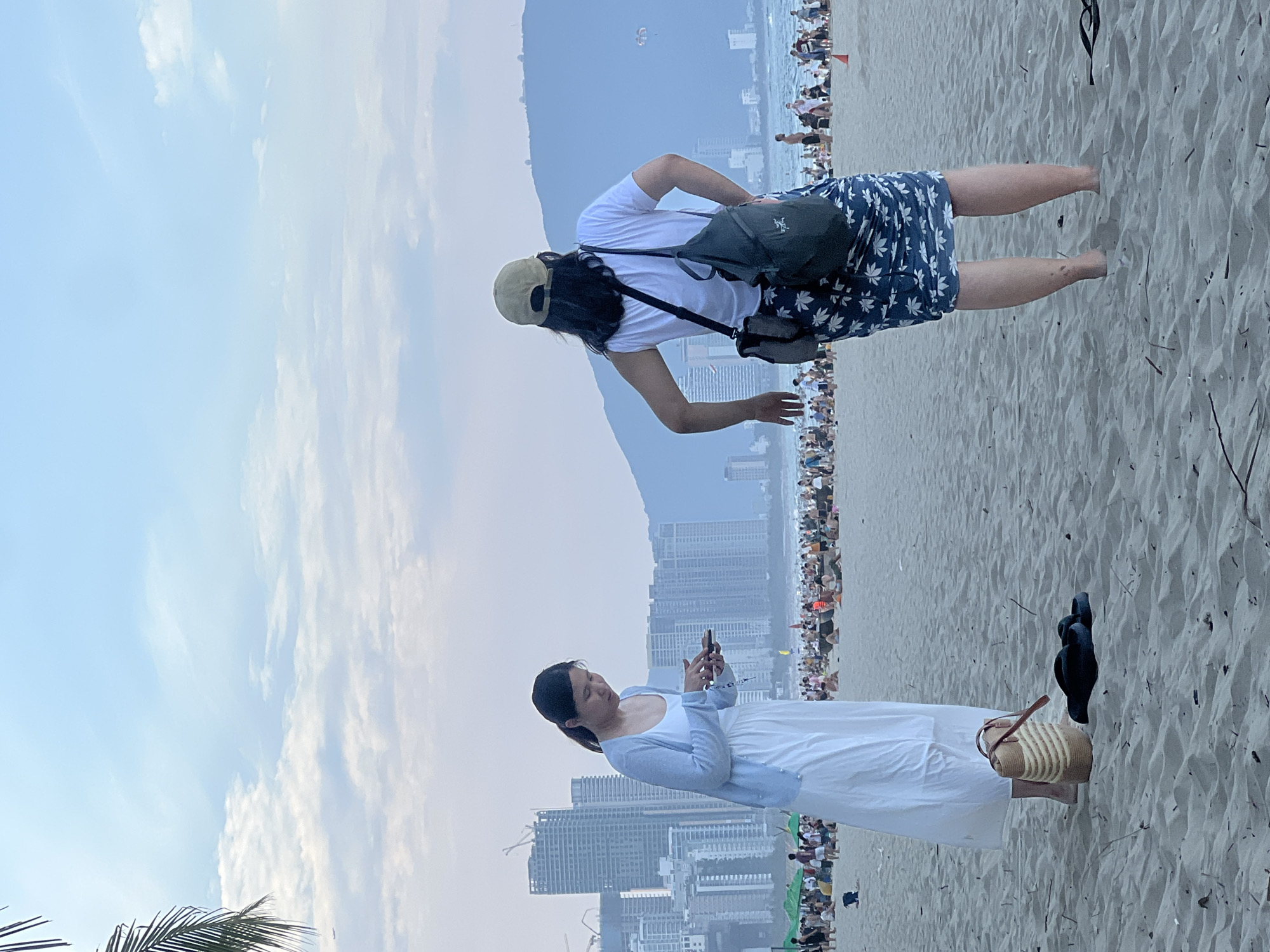 Two individuals are present on a sandy beach in Đà Nẵng, Vietnam, with the cityscape and ocean in the background. The individual on the left, facing away from the camera and towards a smartphone, is wearing a white dress and a light blue cardigan. Next to them, on the sand, is a woven beach bag and a pair of black flip-flops. The second individual, also facing away from the camera, is wearing a white t-shirt, a light brown baseball cap, and a blue skirt with a white floral pattern. They are carrying a dark green backpack and a smaller grey bag. The beach is populated with numerous people, some sitting and others standing. In the distance, the city skyline features several tall buildings, and beyond that, a mountain range is visible under a sky with scattered clouds.