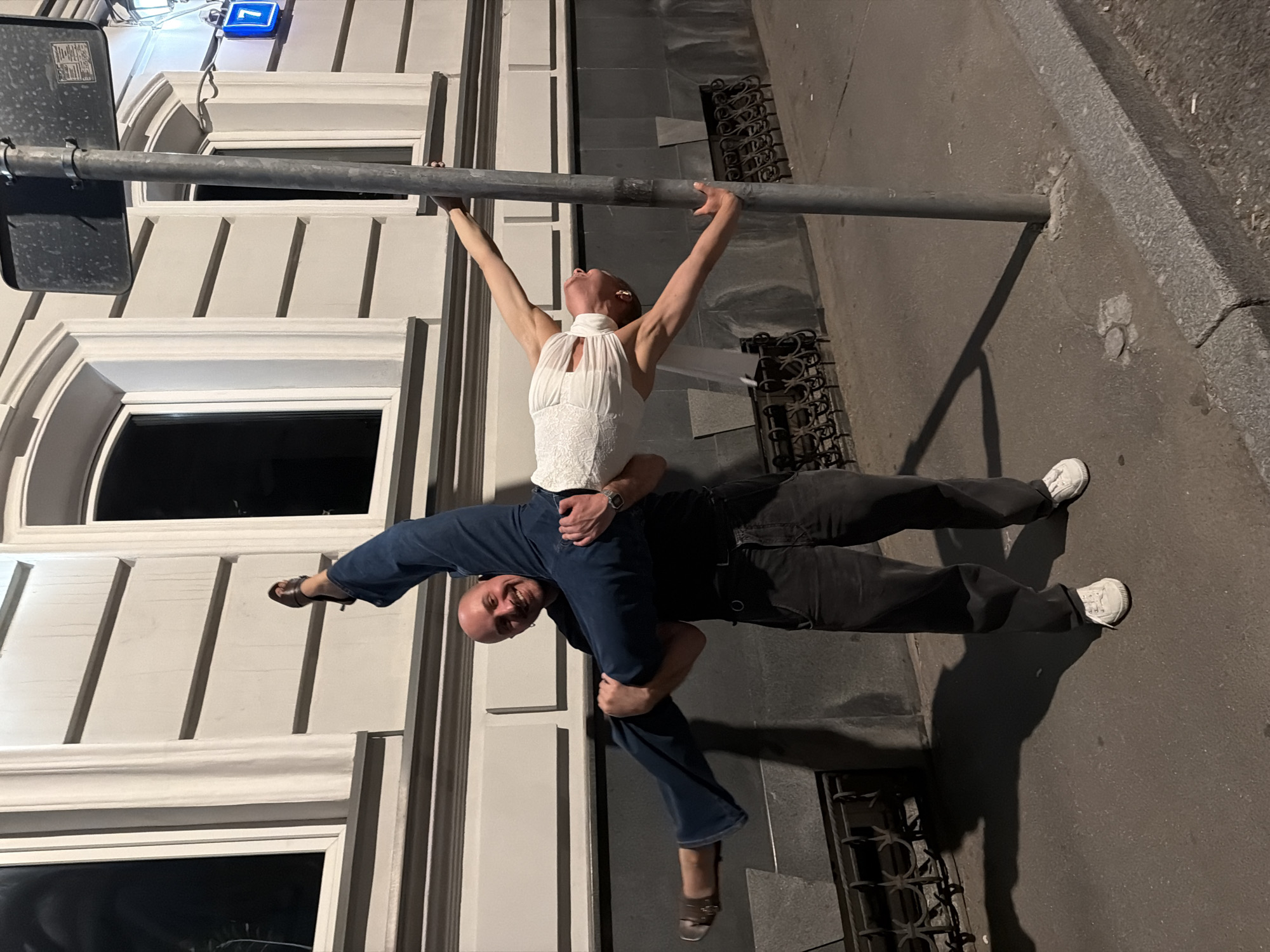 In Moscow, Russia, on a street at night, a bald man in dark pants and white sneakers is holding a woman in a white lace dress. The woman is in a dramatic pose, with her legs extended and her arms outstretched. She is leaning back, with her right hand gripping a metal pole. The man is supporting her with his hands under her legs and back. The building behind them is a light-colored building with dark windows. There is a street light visible in the background, and a blue street number sign with the number 7. The ground is paved with asphalt and there is a concrete curb.