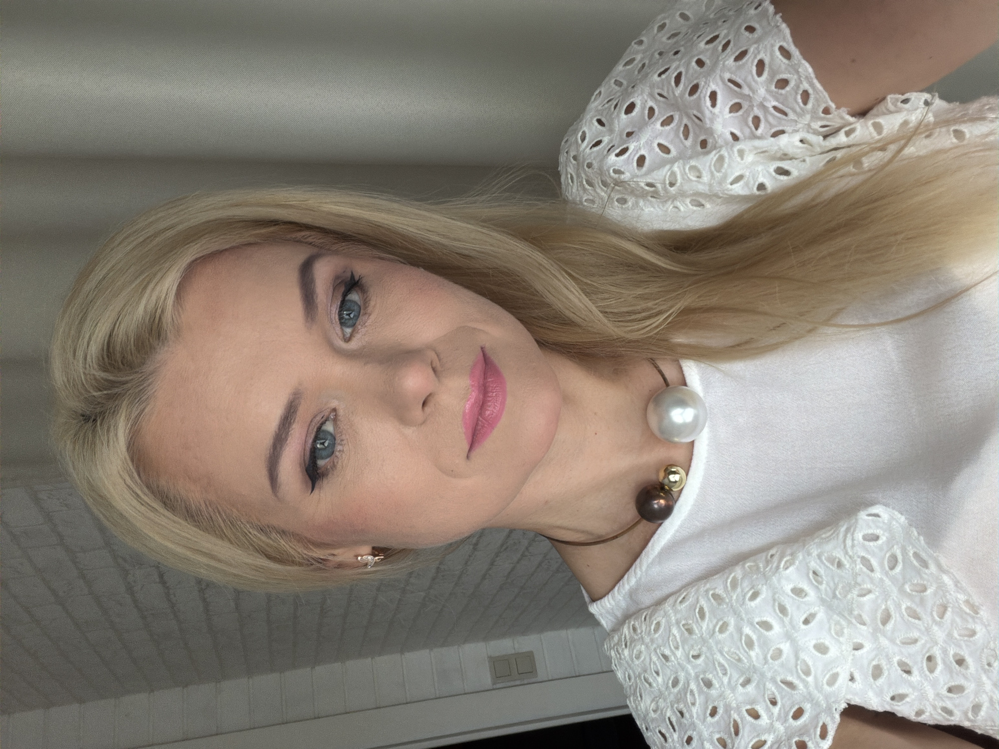 A blonde woman with blue eyes, light skin, and pink lipstick poses for a selfie. She is wearing a white eyelet top and a necklace with three large, round beads in varying shades of brown, gold, and white. The background consists of a white brick wall on the left and a light gray curtain on the right. The woman is indoors, likely in a residential setting. There is no discernible indication of the city of Sochi, Russia, or any urban environment.