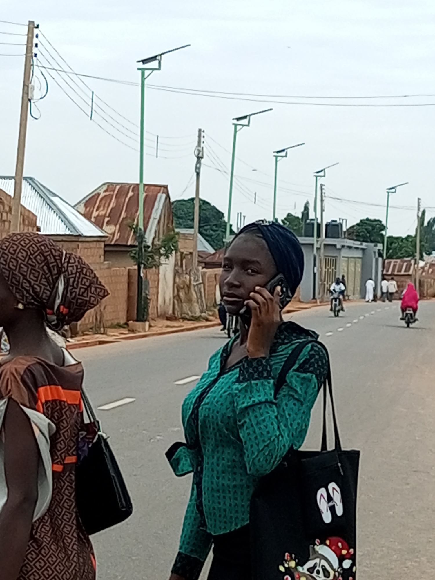 The image depicts a street scene in Gombe, Nigeria. In the foreground, a young woman wearing a blue headscarf and a green patterned top is holding a mobile phone to her ear and speaking. She is carrying a black tote bag with cartoonish imagery. To her left, another young woman is partially visible, wearing a headscarf with a brown and white pattern and a brown and orange dress. Both women appear to be walking on or near a paved road. The background shows a residential area with buildings featuring corrugated metal roofs and brick walls. Several solar-powered streetlights are installed along the road. In the mid-ground, other individuals are seen on motorcycles and walking, suggesting general activity on the street. The sky is overcast.