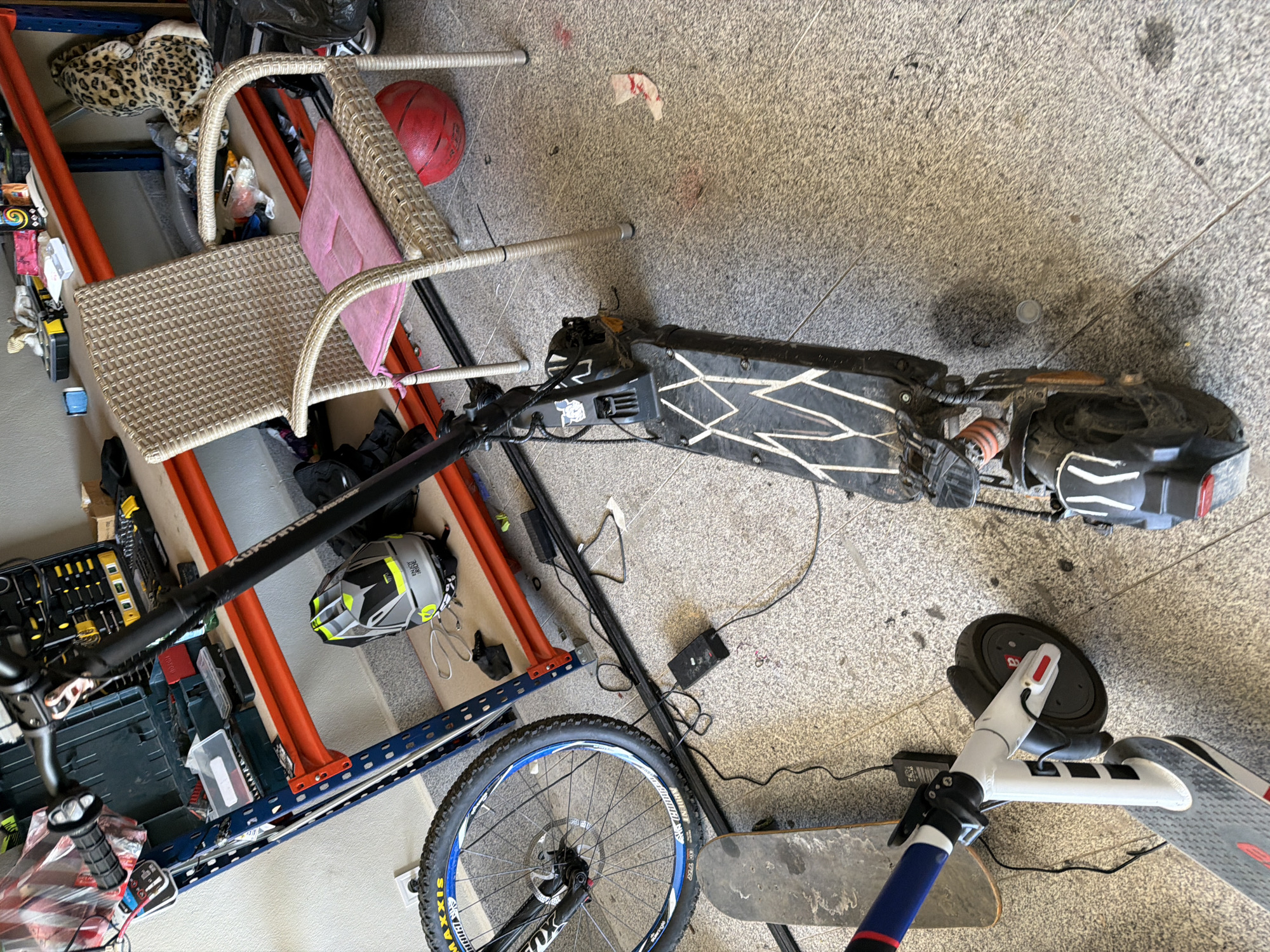 The image shows a cluttered indoor scene, possibly a garage or storage area in Asenovgrad, Bulgaria. A black and white electric scooter lies horizontally on the floor, along with a partially visible bicycle, and another scooter. A rattan chair sits near a shelf unit filled with various items, including tools, containers, and what appears to be a stuffed animal. A helmet is also present on the shelf, adjacent to the bicycle. The scene suggests a storage space with recreational equipment and tools.