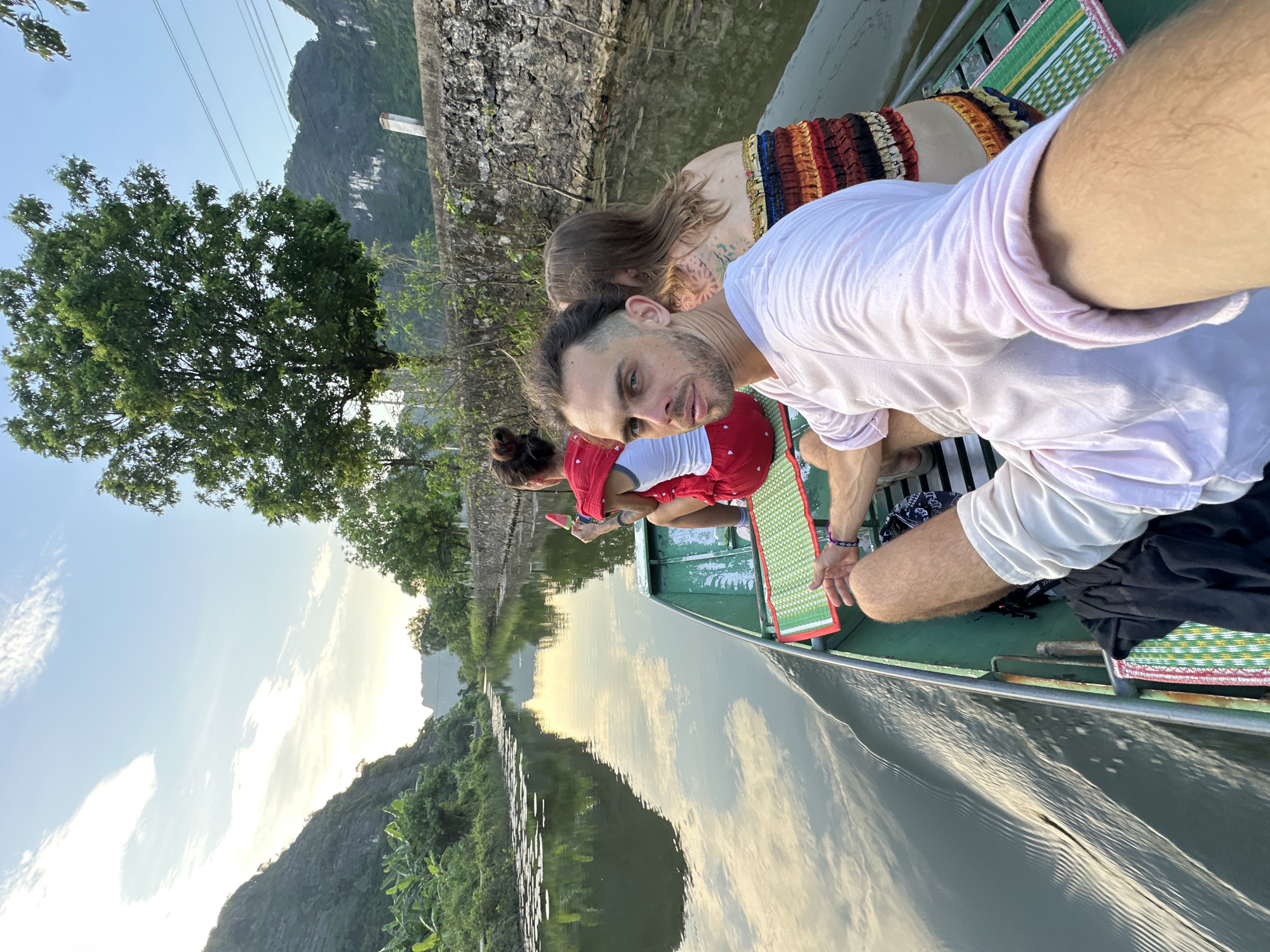 This image captures a serene moment on a boat navigating a calm river in Nam Hoa Lư Ward, Vietnam. The sky is a soft blend of blue and yellow hues, with wispy clouds reflecting on the water's surface, suggesting either sunrise or sunset. Towering, lush green karst mountains rise majestically on either side of the river, their imposing presence softened by the gentle light. Dense vegetation, including banana trees and other foliage, spills down the slopes towards the water. The river itself is dotted with lily pads, adding a touch of natural beauty.
In the foreground, a green boat carries four individuals. The person closest to the camera, a male with short dark hair and a beard, is looking directly at the viewer, his expression neutral. He is wearing a light pink t-shirt and white shorts. To his left, a woman with long blonde hair and a colorful, striped top is visible, her back to the camera. Behind her, another woman, wearing a white tank top and a red skirt with white polka dots, is also facing away from the camera, holding a pink popsicle. The boat is equipped with green mats and a woven mat with green and red trim. The water's surface is disturbed by the boat's movement, creating gentle ripples. The scene is one of peaceful exploration, with the natural landscape of Nam Hoa Lư Ward providing a breathtaking backdrop.