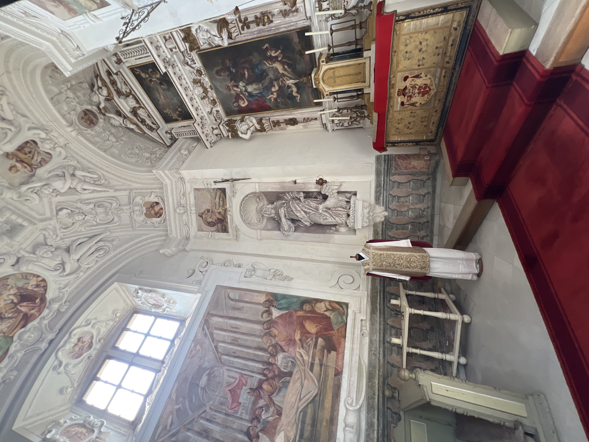The image depicts the interior of a richly decorated church, likely a chapel, featuring ornate stucco work on the vaulted ceiling and walls, adorned with frescoes and paintings. In the foreground, a mannequin is dressed in a white alb and a gold and white chasuble, a liturgical vestment. To the left, a large fresco covers a significant portion of the wall, depicting a scene with numerous figures, a bier, and architectural elements. A wooden table with two candles stands in front of the fresco. To the right, an altar is draped with a red cloth, and behind it, a large ornate tapestry with a coat of arms is displayed. Steps covered in red carpet lead up to the altar. A statue of a saint, possibly John the Baptist given the inscription "IOANNES" visible below it, stands in a niche to the left of the altar. The overall impression is one of solemnity and historical grandeur. This scene is located in the Náchod Castle in Náchod, Czechia, specifically within the Castle Church of Saint Peter and Saint Paul.