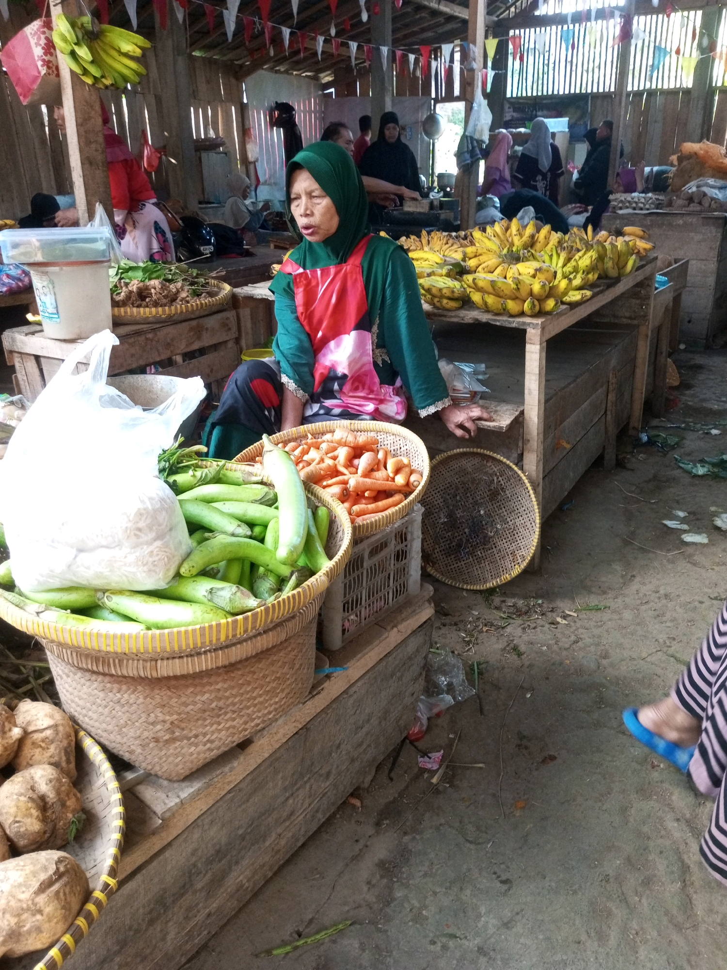 In Kalibeber, Indonesia, a woman wearing a green hijab and a red apron stands behind a stall filled with fresh produce. She is looking upwards with her mouth slightly open. In front of her, a large woven basket overflows with green eggplants, and a white plastic bag containing what appears to be noodles or shredded vegetables sits atop them. To her right, another woven basket holds a pile of bright orange carrots. Behind her, several bunches of yellow bananas are displayed on a wooden table. Other people, some wearing hijabs, are visible in the background, suggesting a busy market atmosphere. The stall is constructed of wood, with a dirt floor and overhead lighting. The overall scene depicts a typical market environment in a rural Indonesian setting.