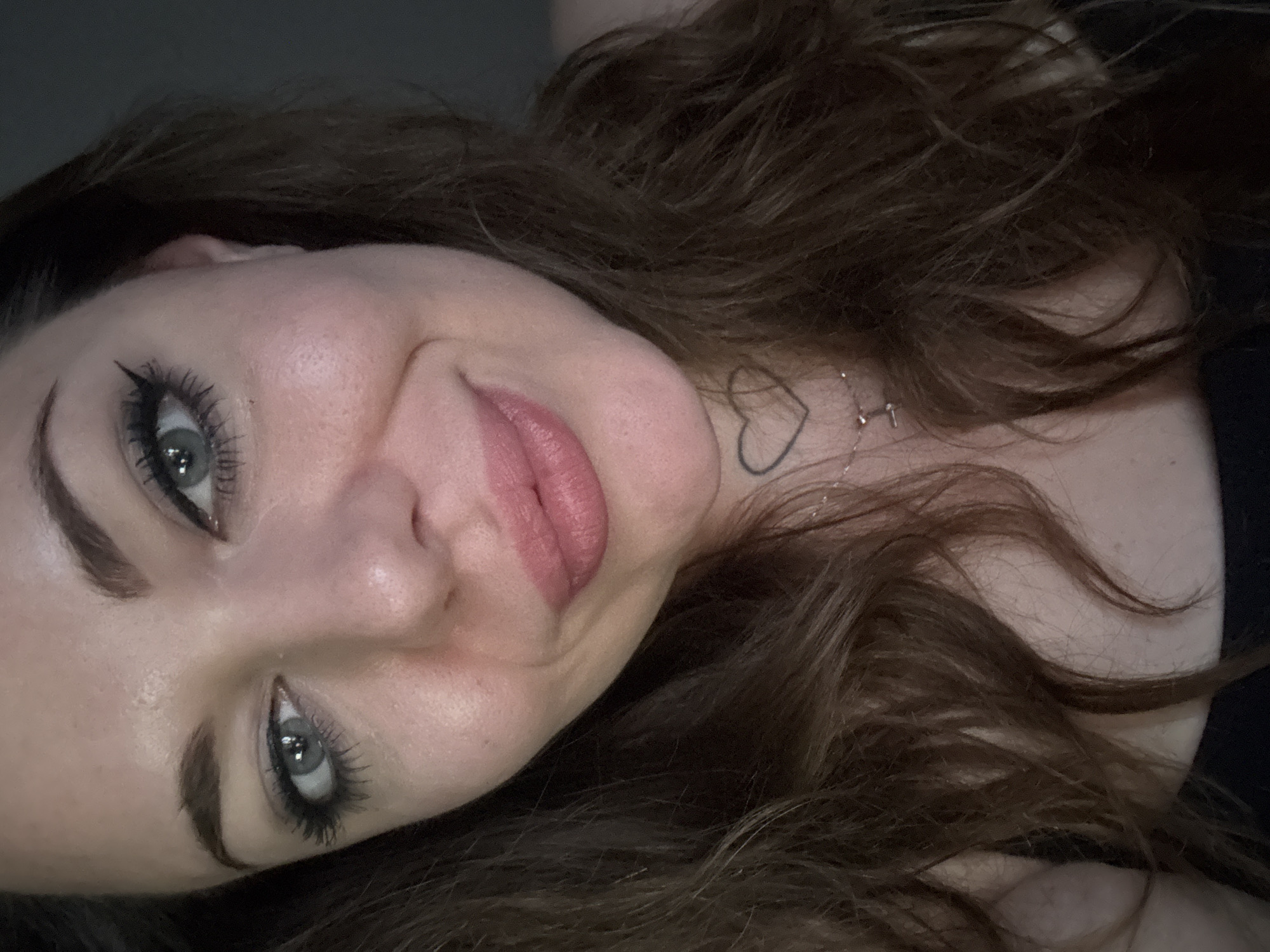 The image is a close-up, head-and-shoulders selfie of a young woman with brown, wavy hair. She has fair skin, light-colored eyes with dramatic winged eyeliner and mascara, and pink lipstick. A small heart tattoo is visible on her neck. She is wearing a black top. The background is a dark, plain surface, likely a wall or curtain, and does not provide any identifiable details of a specific location, including the city of Kremenchuk, Ukraine.
