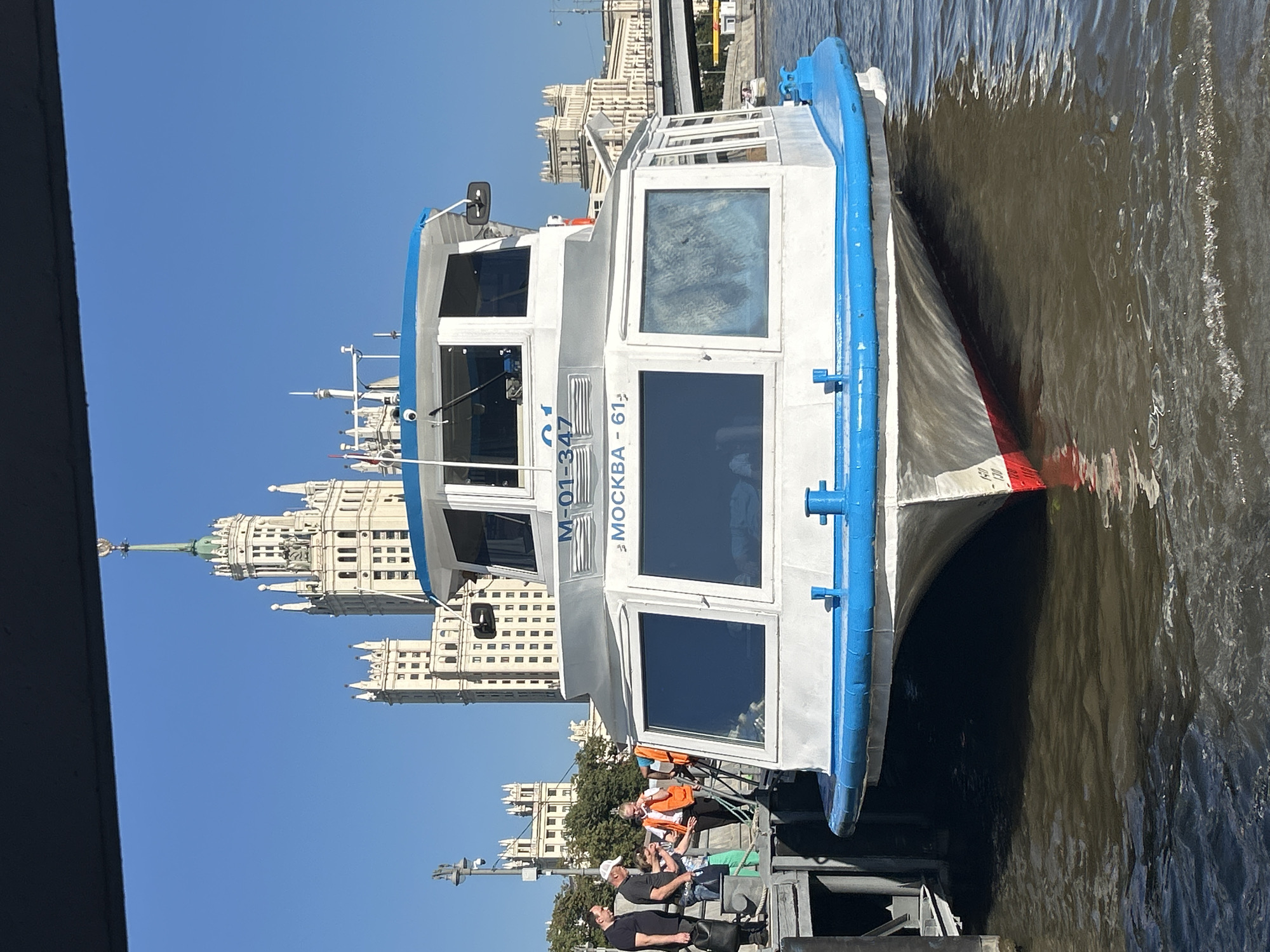 The image shows a white passenger boat with a blue trim docked on a river in Moscow, Russia. The boat has large windows and a cabin on top. Several people are visible on the dock near the boat, some appearing to be passengers and others potentially crew members wearing orange vests. In the background, iconic Moscow architecture can be seen, including a tall, ornate building with spires that resembles the Stalinist skyscrapers. A bridge and other buildings are also visible in the distance, suggesting an urban riverside setting. The sky is clear and blue, indicating a sunny day. The water in the river appears calm with some ripples.