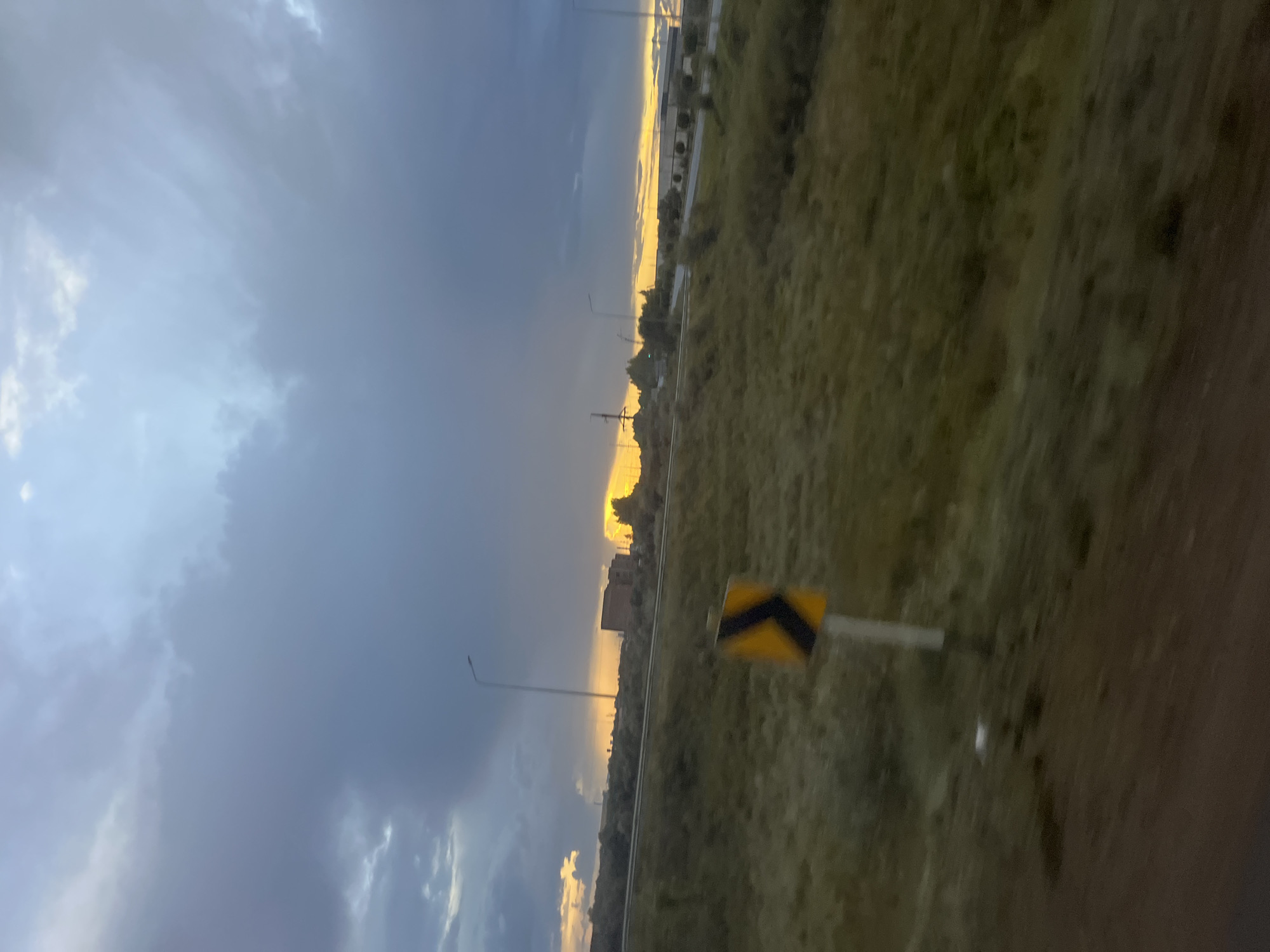 This image depicts a road scene in Astana, Kazakhstan, during sunset. The sky is dominated by dramatic, dark clouds, with a bright orange and yellow glow emanating from the horizon, indicating the setting sun. In the middle ground, there is a road running horizontally across the frame, bordered by vegetation. A yellow and black directional arrow sign is visible on the right side of the road. Beyond the road, there are some industrial buildings and trees. The foreground shows a patch of dark soil and blurred green vegetation, suggesting the photo was taken from a moving vehicle. The overall impression is one of a somewhat overcast sunset over a semi-industrial landscape.