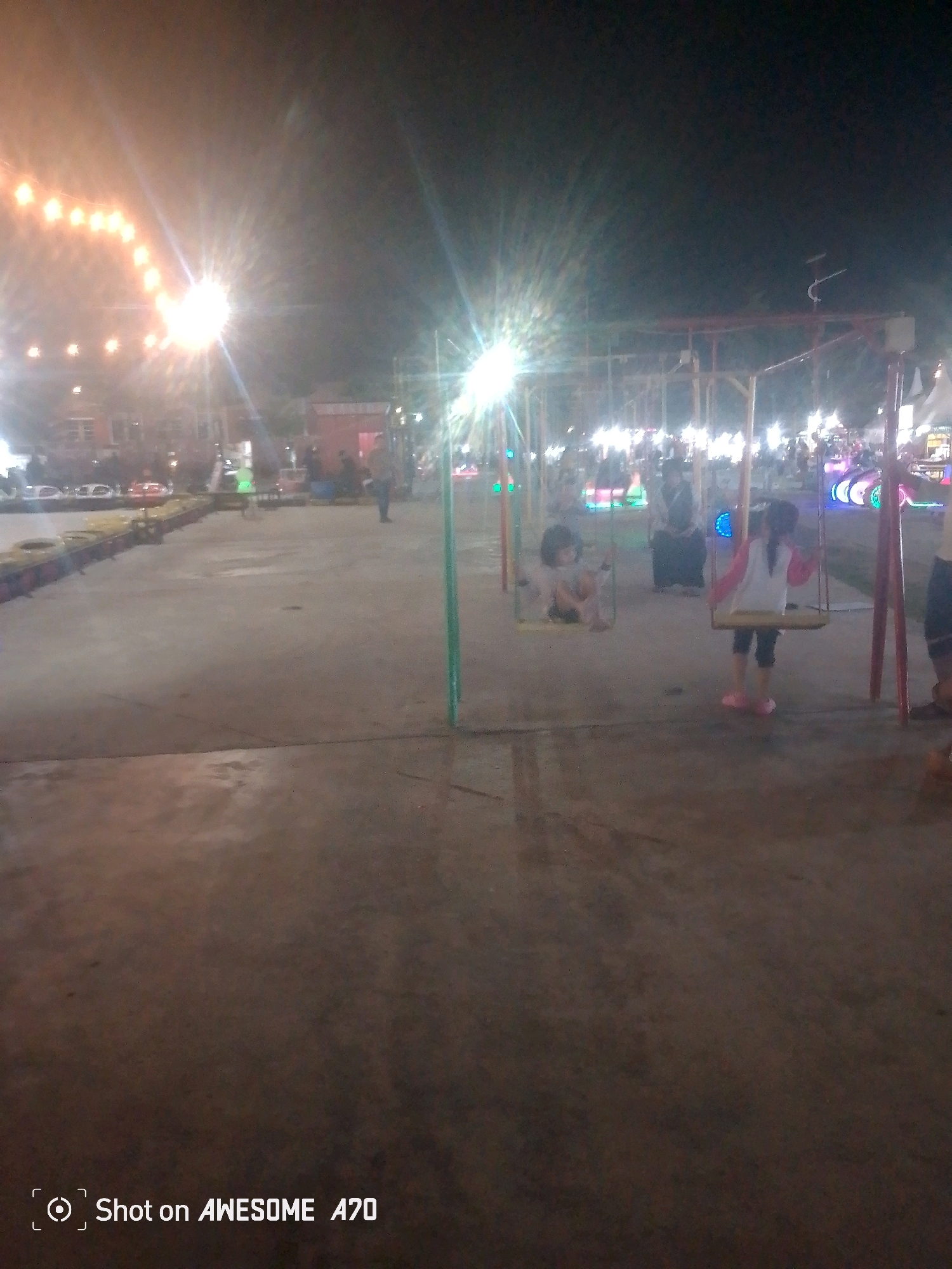 An outdoor scene at night, possibly an amusement park or a night market, is depicted. The ground appears to be concrete. In the foreground, there are the faint outlines of what could be a go-kart track. The middle ground features children on swings, with one child sitting and another standing. In the background, there are various structures, lights, and a crowd of people. The overall lighting is dim, with bright lights from strings of bulbs and other illumination sources creating lens flares and a slightly blurry effect. The image is stamped "Shot on AWESOME A70". The location is identified as Batam, Indonesia.