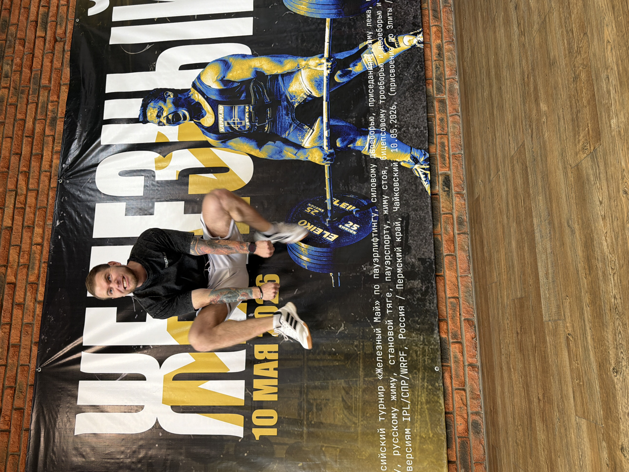 The image shows a man lying on a promotional banner, with his body oriented perpendicular to the long edge of the banner. The banner is laid out on the floor, bordered by a brick wall on one side and wood flooring on the other. The man is Caucasian, has short brown hair, a visible beard, and tattoos on his arms. He is wearing a dark grey t-shirt, light-colored shorts, and white Adidas shoes with black stripes. He is smiling and looking at the camera.
The banner's design includes a weightlifter illustration in yellow and blue tones, and it has the words "ЖЕЛЕЗНЫЙ" in large letters. It also lists information about a powerlifting tournament called "Iron May," including the date (May 10) and the location in Chaikovsky, Russia. The banner includes a circular "ELEIKO" logo with the text "25 SWEDEN."