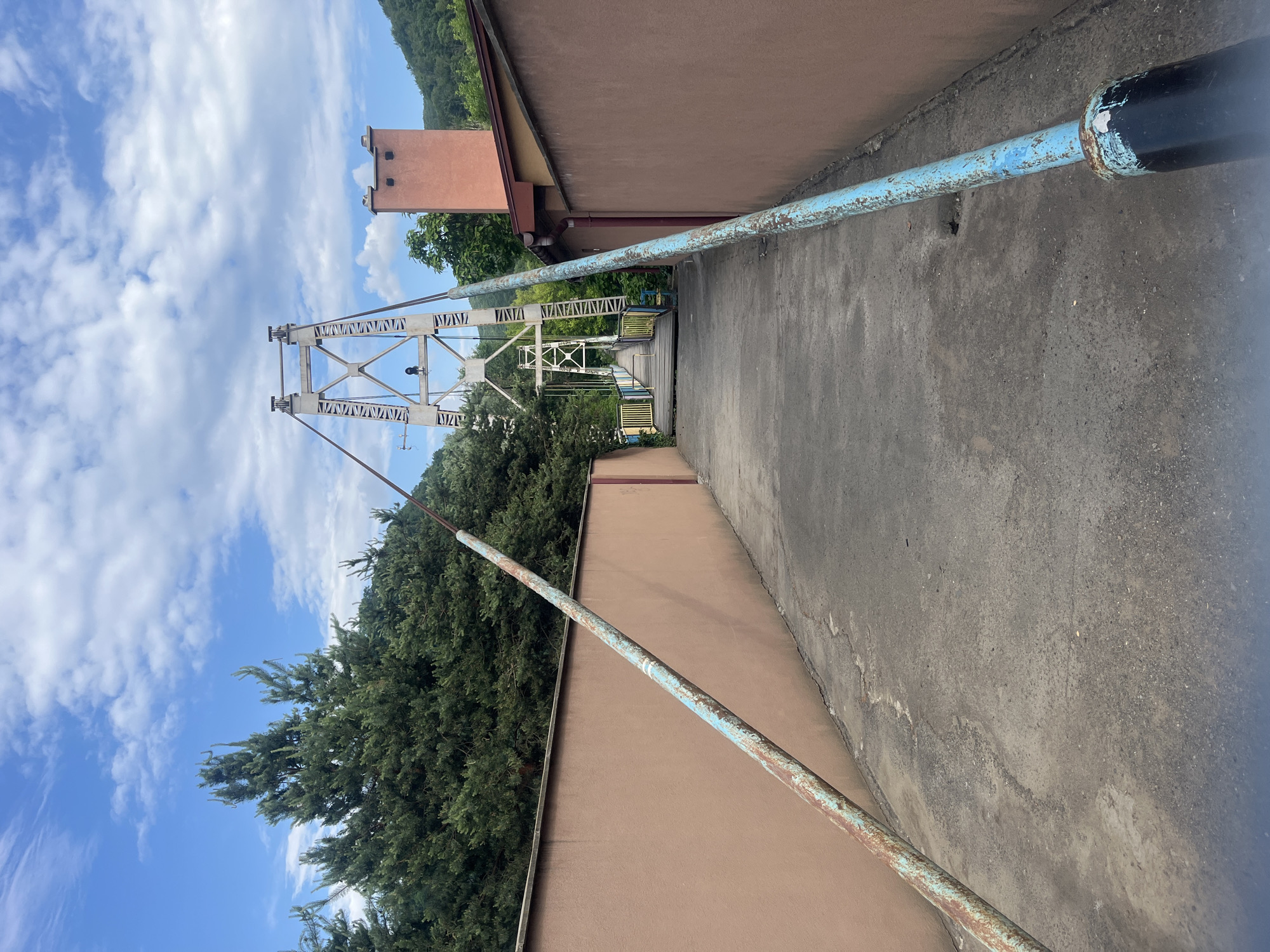 An outdoor scene captured in Kamianytsia, Ukraine, features a pedestrian suspension bridge stretching across the center of the frame, viewed from an elevated paved pathway. The bridge is constructed with distinctive A-frame metal towers and a truss-like structure painted white or light grey, showing signs of weathering. On the vertical members of the bridge's framework, the word "ZAKARPATTYA" is visibly repeated in white text. The bridge deck appears to be made of wood or a similar material, with visible handrails.
To the right of the paved pathway, a building with a light brown/tan wall runs parallel to the path. A rusty, light blue metal pipe or railing extends along the edge of this path, separating it from the building. In the distance, behind the bridge and to the left, another building with a reddish-brown section and a chimney-like structure is visible.
The surrounding environment is lush with greenery, including dense conifer trees and bushes, particularly on the left side of the frame and around the base of the bridge. Rolling green hills covered in dense foliage can be seen in the far background. The sky is bright blue with scattered white clouds, indicating clear weather and daytime. No people or specific activities are depicted in the image, suggesting a tranquil and static scene.