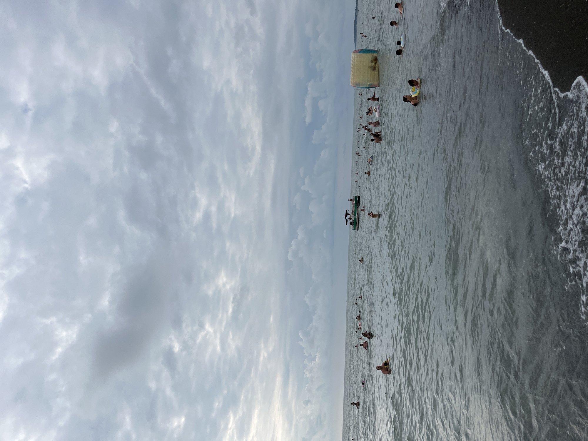 The image captures a crowded beach scene in Ureki, Georgia, under a cloudy sky. The sea is filled with numerous people swimming, wading, and enjoying various water activities. In the mid-ground, a pontoon boat with several individuals on board is visible. To the right, a large inflatable water toy, possibly a hamster ball or roller, is floating on the water, with people interacting with it. The foreground shows the dark sand of the beach with gentle waves lapping at the shore. The overall atmosphere is one of a busy summer day at a popular seaside resort.