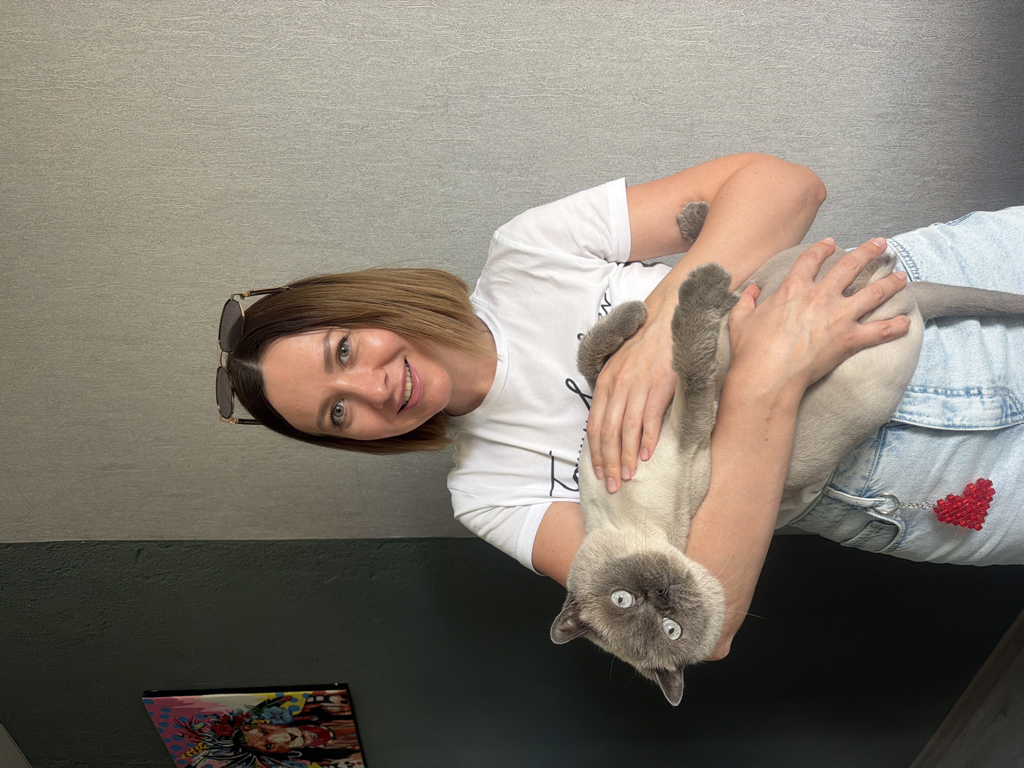 In an indoor setting, a woman with shoulder-length brown hair and sunglasses perched on her head is shown holding a light-colored cat. The woman smiles warmly, looking directly at the camera. She is wearing a white t-shirt with some indistinct black lettering and light blue denim jeans, from which a vibrant red beaded heart keychain hangs. The cat, which appears to be a Siamese or similar breed with distinctive blue eyes and light grey/tan fur with darker points on its face, ears, paws, and tail, is also looking towards the camera, seemingly calm and comfortable in her arms.
The background consists of two distinct wall sections: an upper part with a textured, light grey wallpaper, and a lower part with a darker, plain wall. Partially visible in the bottom left corner is a colorful, possibly framed artwork or picture, featuring a graphic, pop-art style illustration, though no specific text on it is legible. The scene depicts a casual and affectionate interaction between the woman and her pet, likely captured in Rostov-on-Don, Russia. The lighting is bright and even, characteristic of an indoor environment.