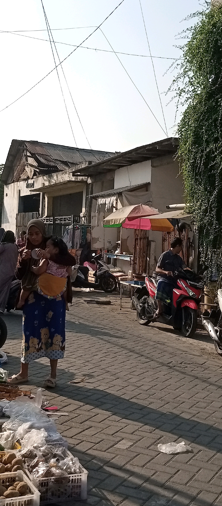 In Wadungasri, Indonesia, a woman wearing a hijab and a patterned blue skirt carrying a baby on her back walks on a brick-paved street. The child, wearing a pink and white patterned outfit, is drinking from a bottle. They are in front of a dilapidated building with a corrugated metal roof. To the right, a man on a red motorcycle is looking towards the left. In the background, more motorcycles and stalls with various items, including what appear to be vegetables, are visible. The scene is illuminated by sunlight, casting long shadows. Overhead, power lines crisscross the sky, and green foliage from a vine-covered structure is visible on the far right.