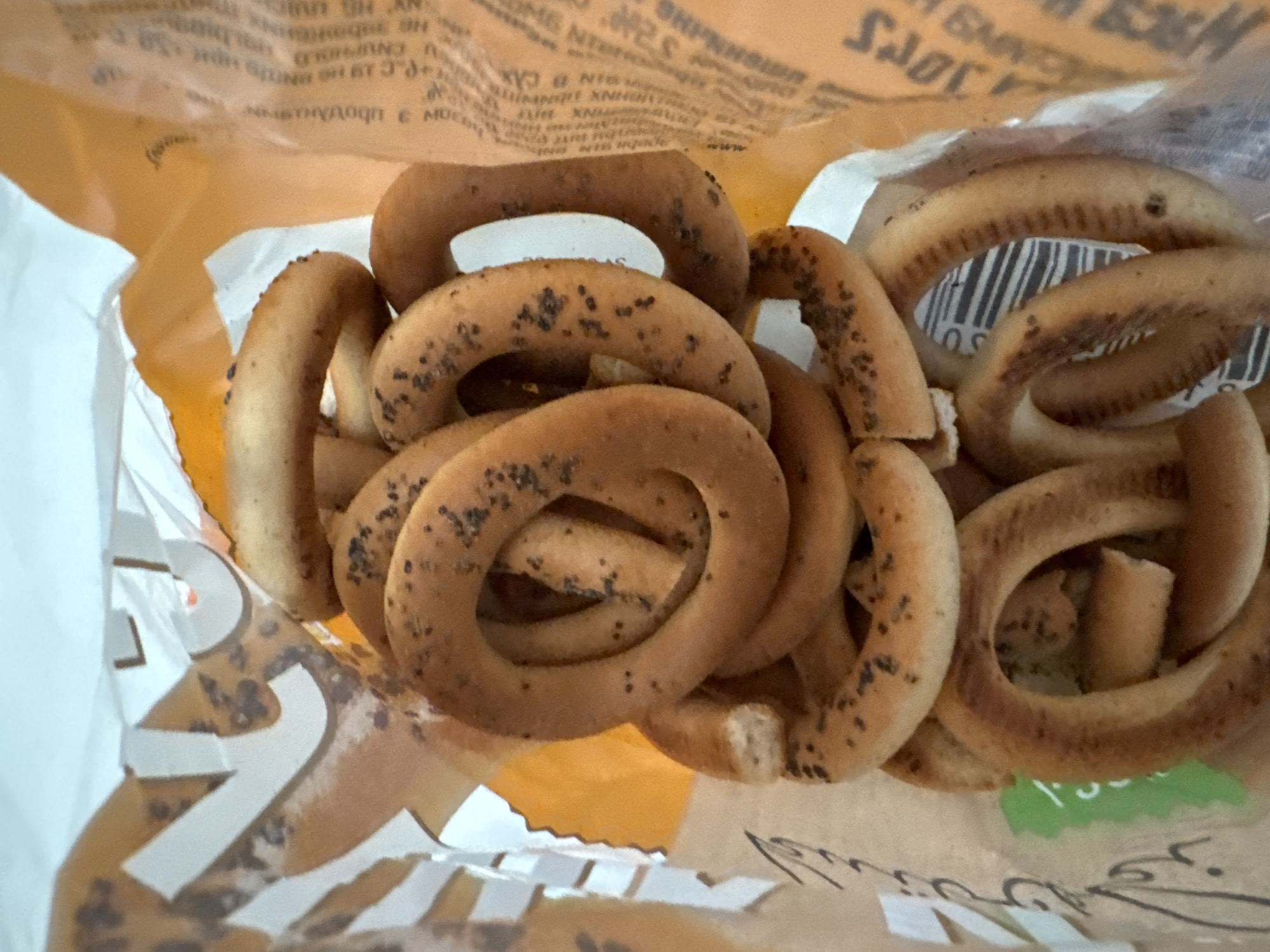 A close-up, top-down shot displays a pile of dried bread rings, possibly bagels or bubliki, inside a white and orange plastic bag. The bread rings are a golden brown color and are speckled with poppy seeds. Some of the rings are broken or cracked. The bag is partially open, revealing more rings stacked within. The background is out of focus, but appears to be a light-colored surface, possibly a table or floor. The location is the Agios Tychona Community, Cyprus. The image is a still life with no human presence or activity.