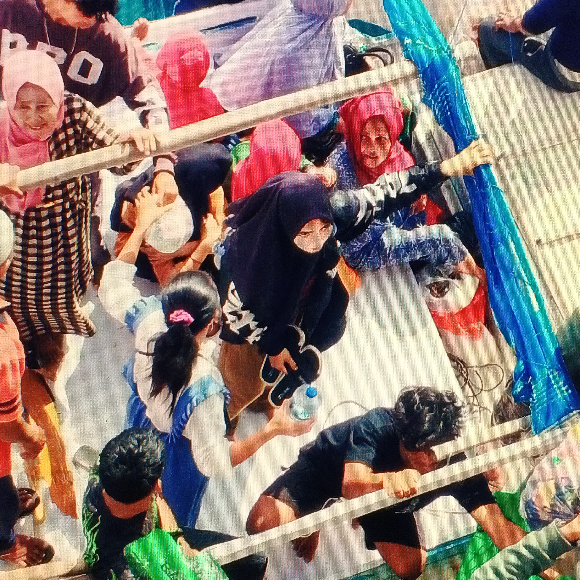 This is a high-angle, slightly blurred, medium shot of a group of people on a boat, likely in the Kelimutu area of Indonesia. The people are dressed in casual clothing, and many are wearing headscarves or hijabs in various colors including pink, red, and black. One person in the foreground, wearing a black hijab and a patterned top, is holding a water bottle and what appear to be sandals. Another person in the mid-ground, wearing a pink hijab and a checkered shirt, is looking towards the viewer. In the background, other individuals are visible, some obscured by the boat's railing and other passengers. A bright blue netting is draped along the side of the boat. The overall scene suggests a crowded, possibly tourist-related, activity on the water.