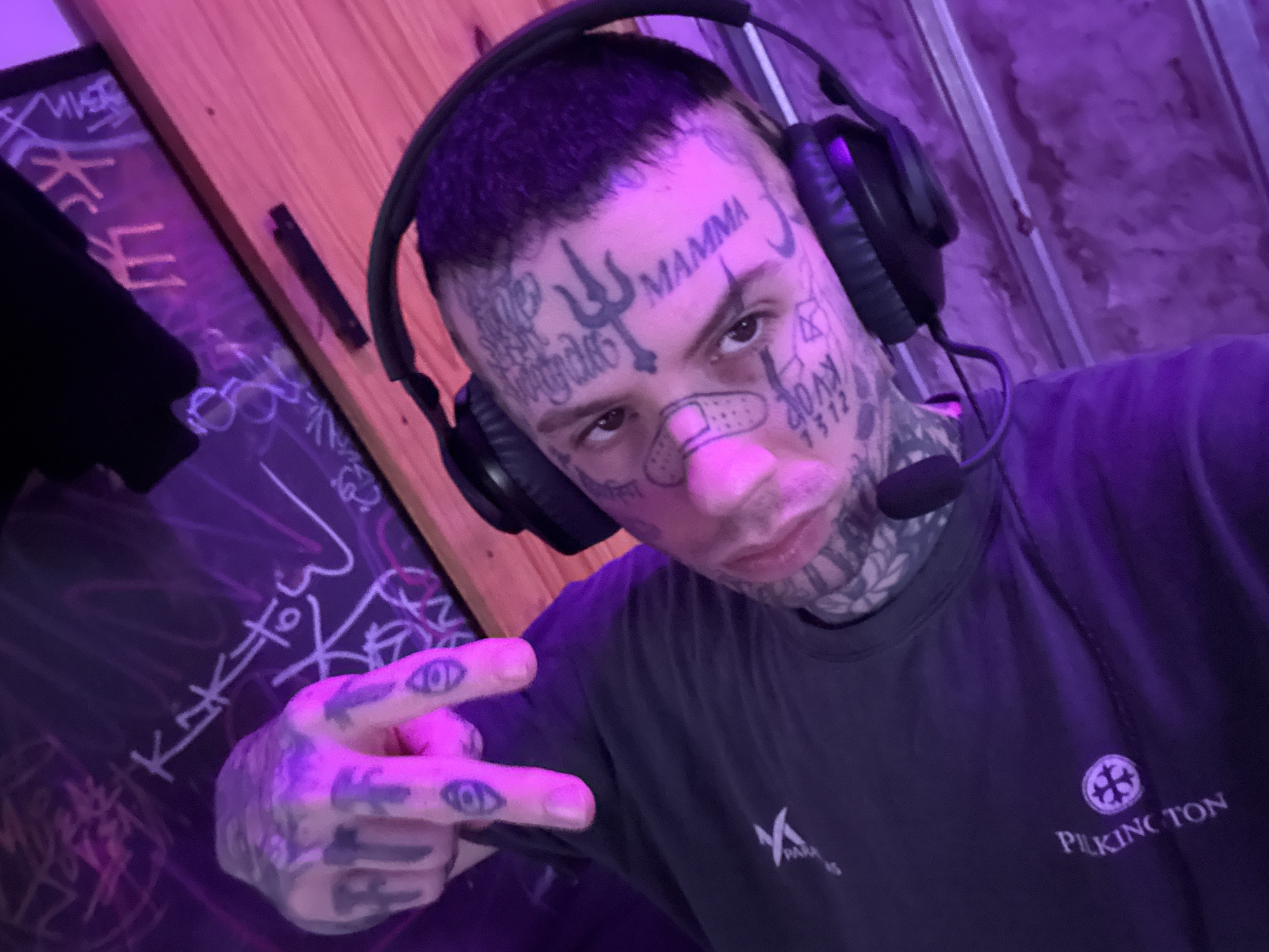 A selfie of a person with extensive facial and hand tattoos is presented. The person is wearing a headset with a microphone and a dark t-shirt with the word "PILKINGTON" and a logo visible on the front. They are making a hand gesture. A bandage is on the nose. Background is dimly lit and contains a wall and a dark board covered in writing. The location in Buenos Aires, Argentina, is not indicated in the image.