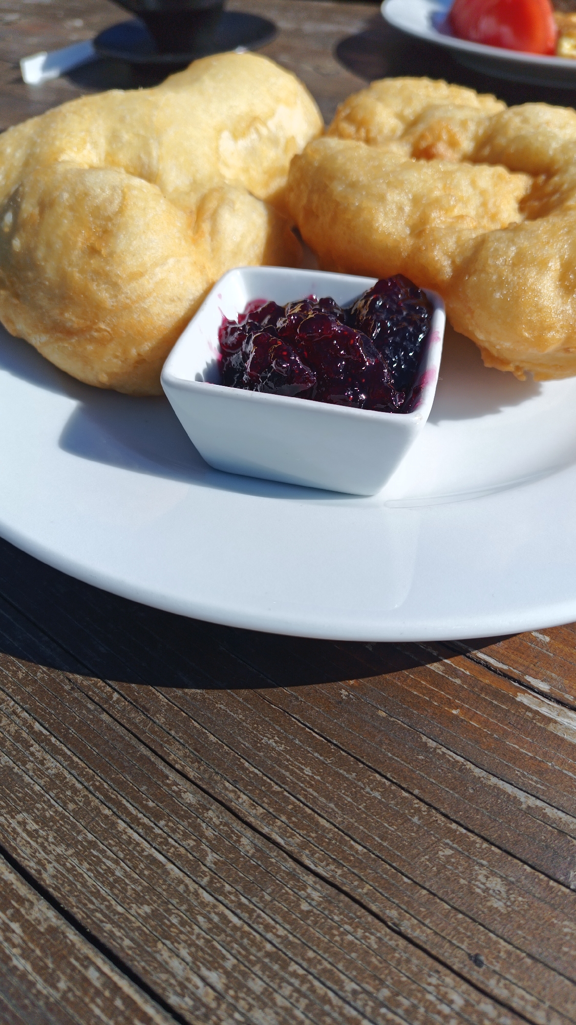A close-up shows a plate of mekitsa, a type of fried dough pastry, with a small dish of berry jam on a wooden table. The scene is located in the city of Vrata, Bulgaria. The mekitsa is golden brown and puffy, while the jam is a deep purple color. The plate is white and the table is made of weathered wood planks.