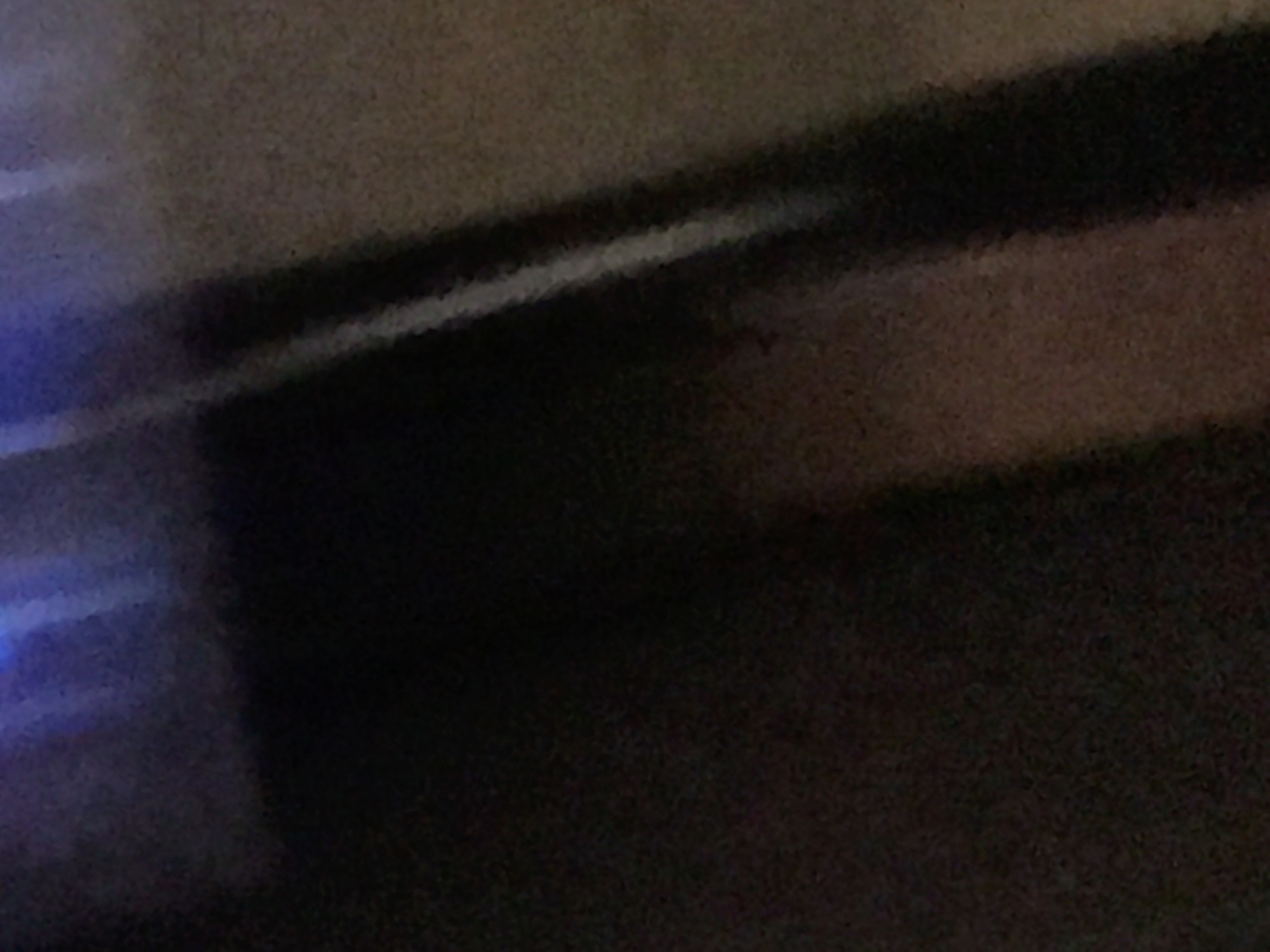 The image is dark and blurry, making it difficult to discern details. There's a diffuse blue light source in the lower-left corner. A lighter, grainy surface stretches across the top, contrasting with a dark, seemingly solid area below. There's a horizontal, lighter line separating the two. No clear indicators of location (Yerevan, Armenia) are visible. No people, subjects, objects, actions, or interactions are discernible. The environment is ambiguous due to the poor image quality.